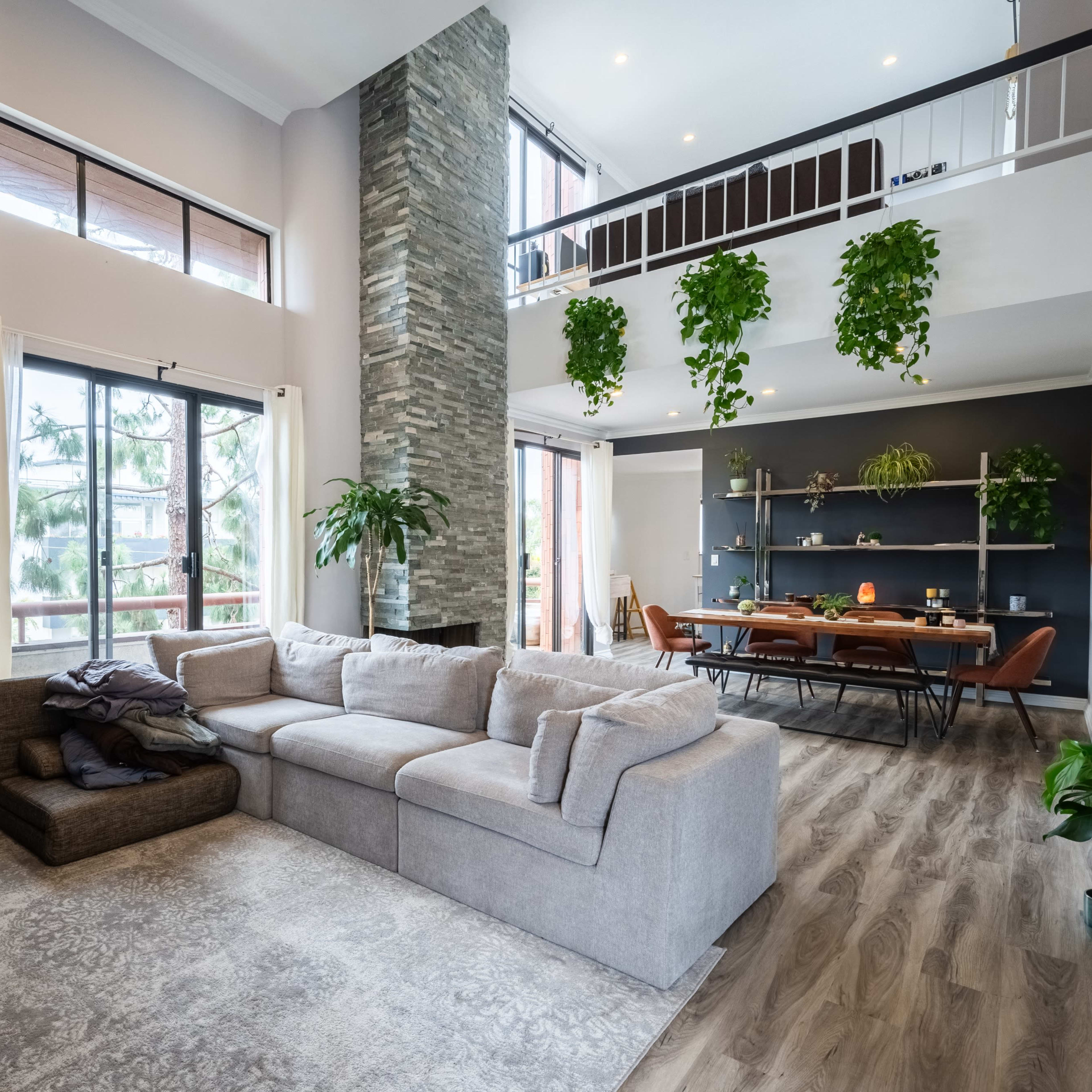 A spacious living room with a large sectional sofa, tall windows, and a dining area with a wooden table and plants hanging from the ceiling.
