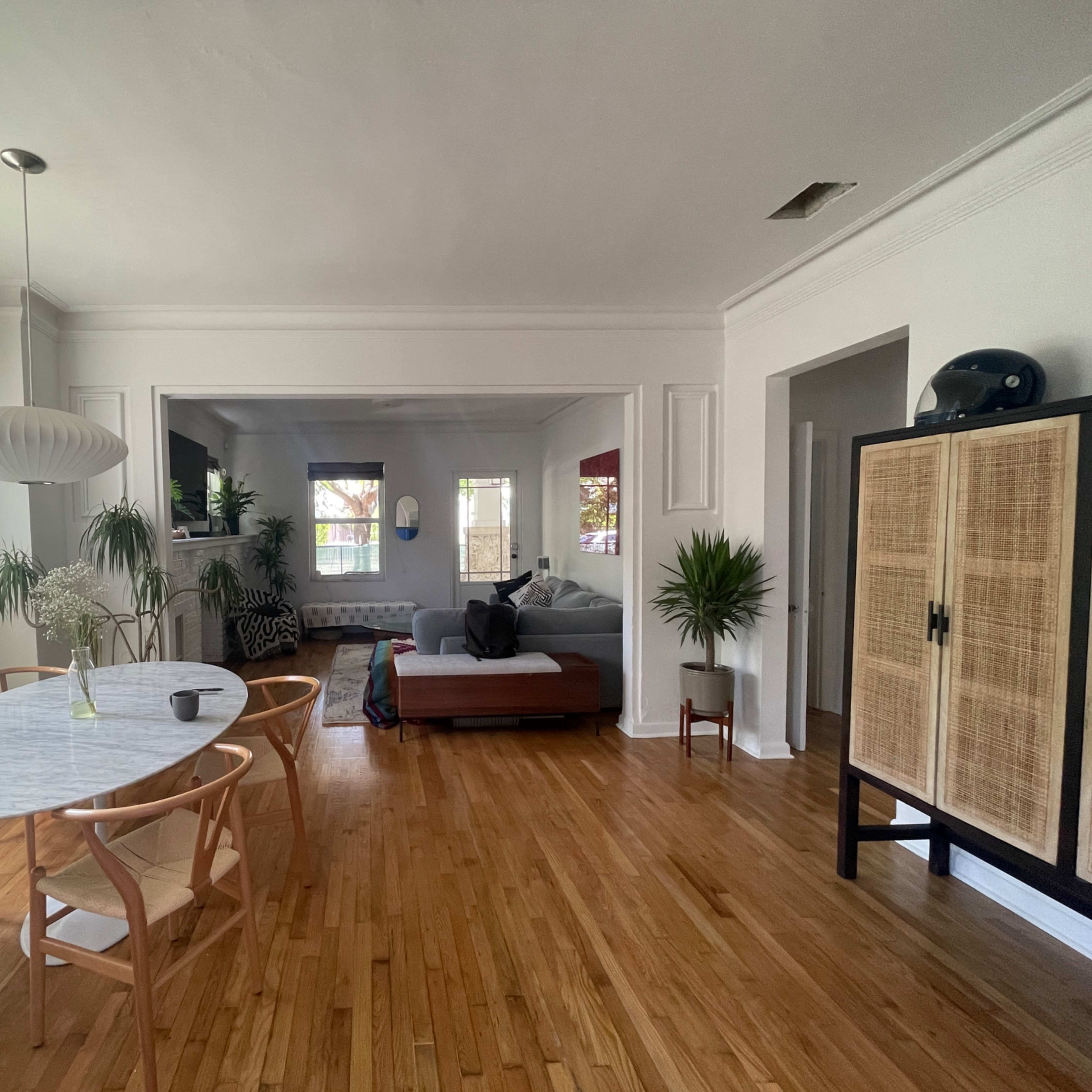 A spacious living area with a dining table and chairs on one side, a sofa in the adjacent room, and large windows allowing natural light to enter.