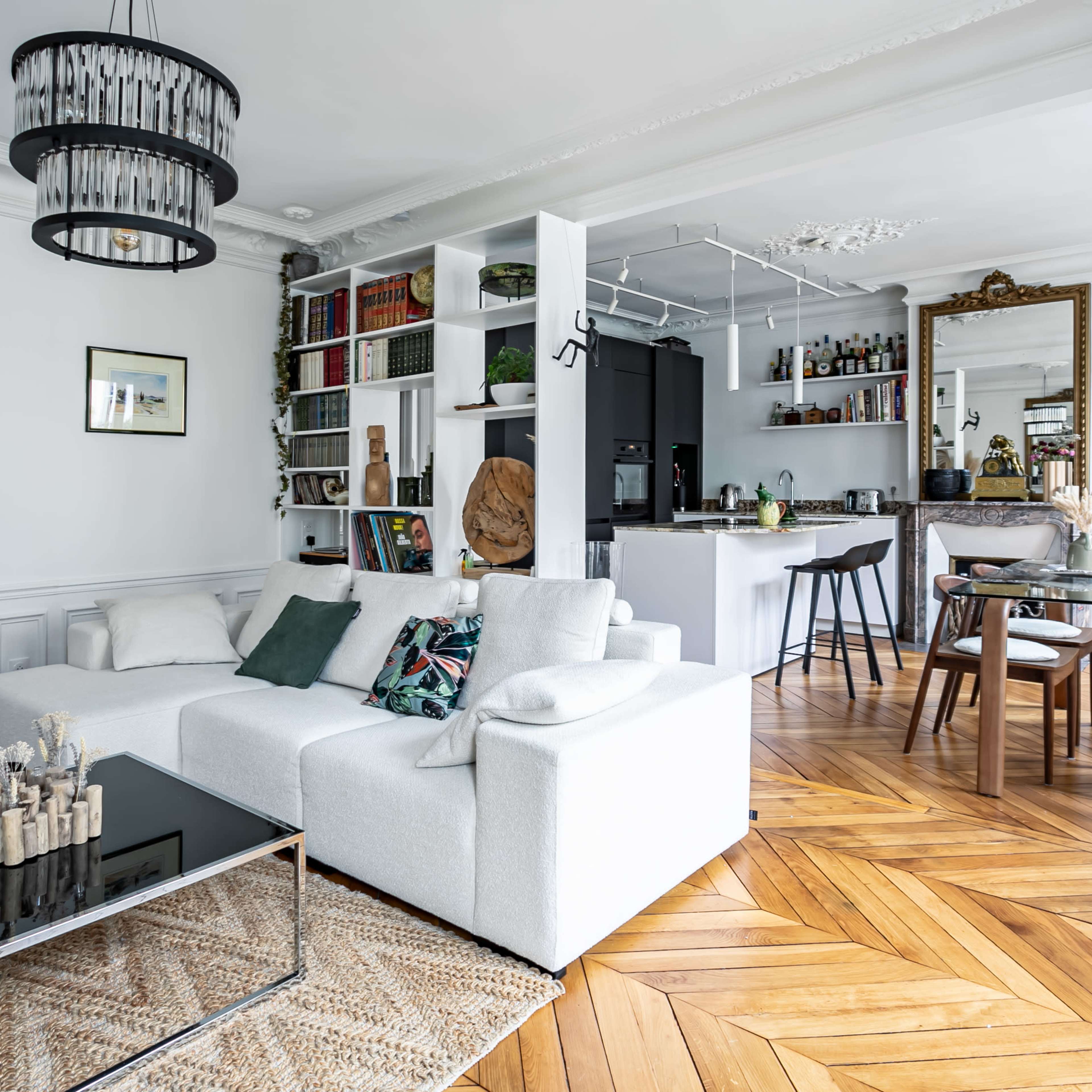 A modern living room with a white sectional sofa, a glass coffee table, and an open kitchen-dining area with stylish decor and wooden flooring.