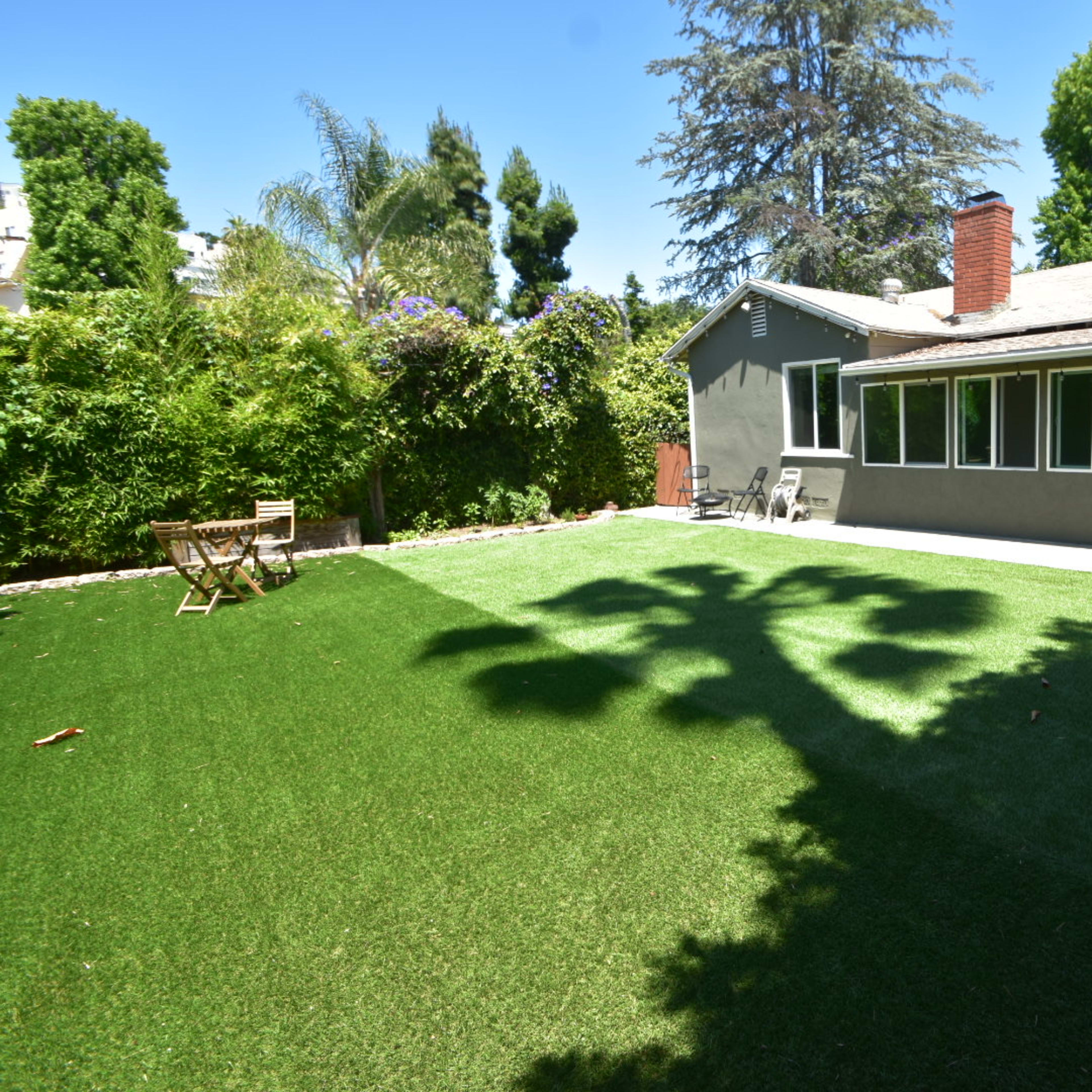 A backyard with artificial grass, featuring a patio area and a few chairs, surrounded by greenery and trees.