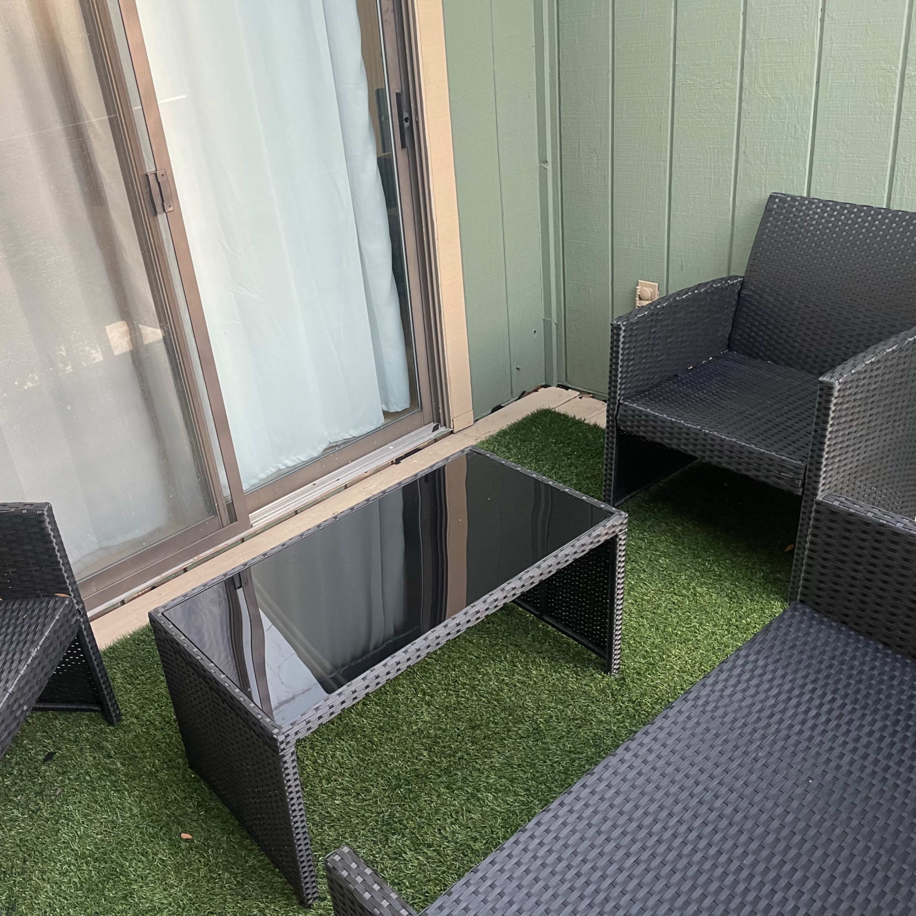 A small outdoor seating area features four black wicker chairs arranged around a glass-top table on green artificial grass.