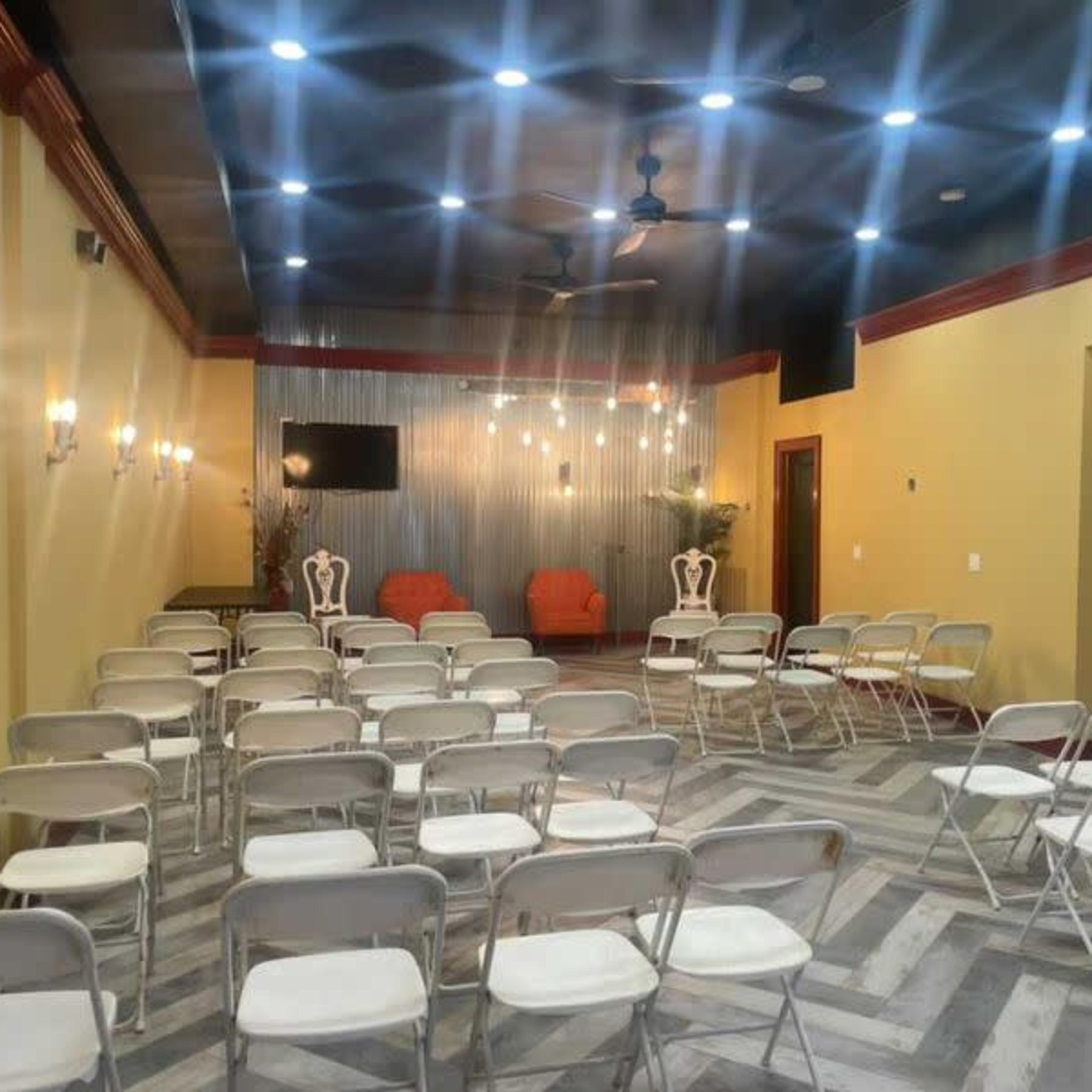 The room features rows of folding chairs facing a decorative wall with two orange chairs and a wall-mounted television, illuminated by overhead lights and sconces.