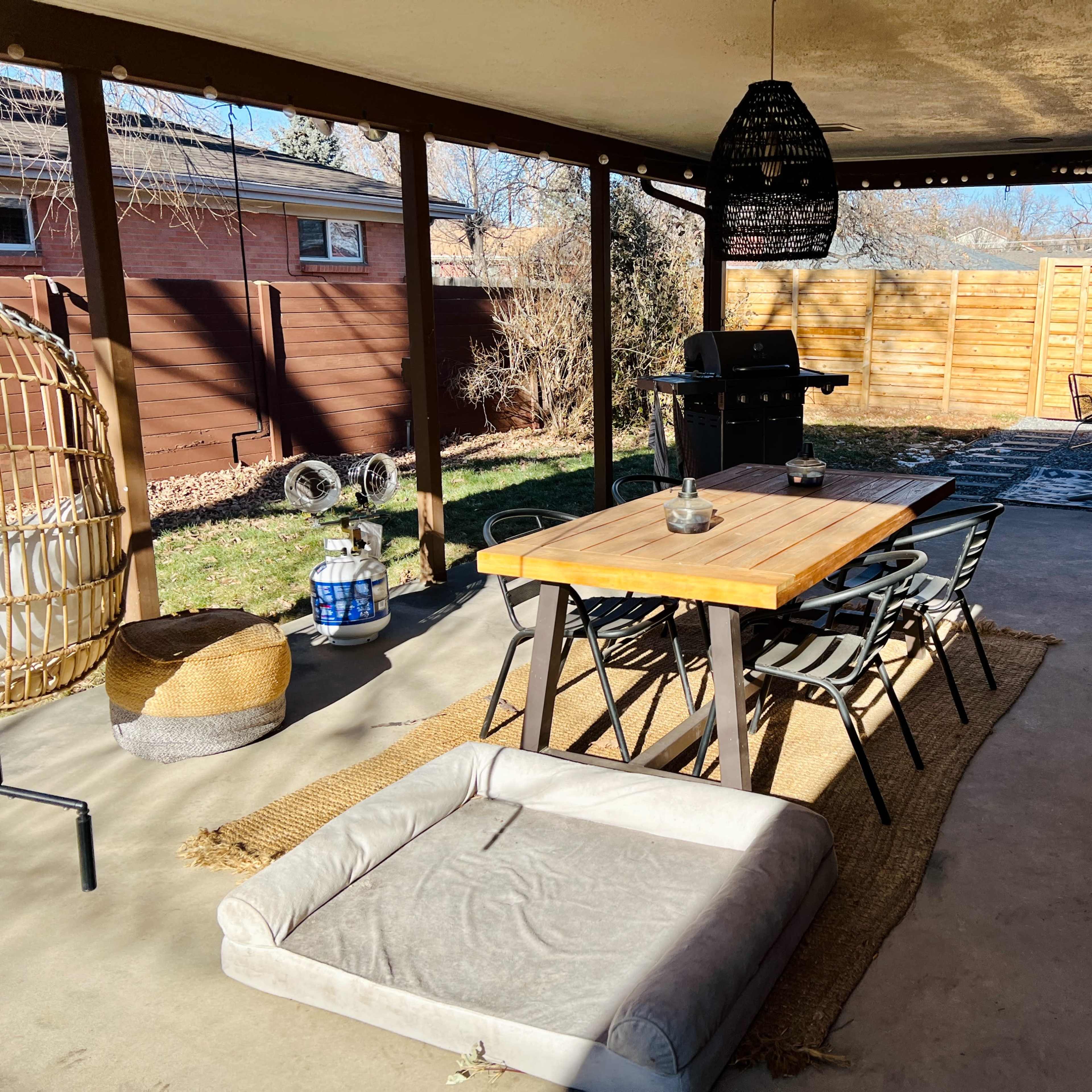 A covered outdoor patio area with a wooden dining table, chairs, a grill, and a dog bed on a concrete floor.