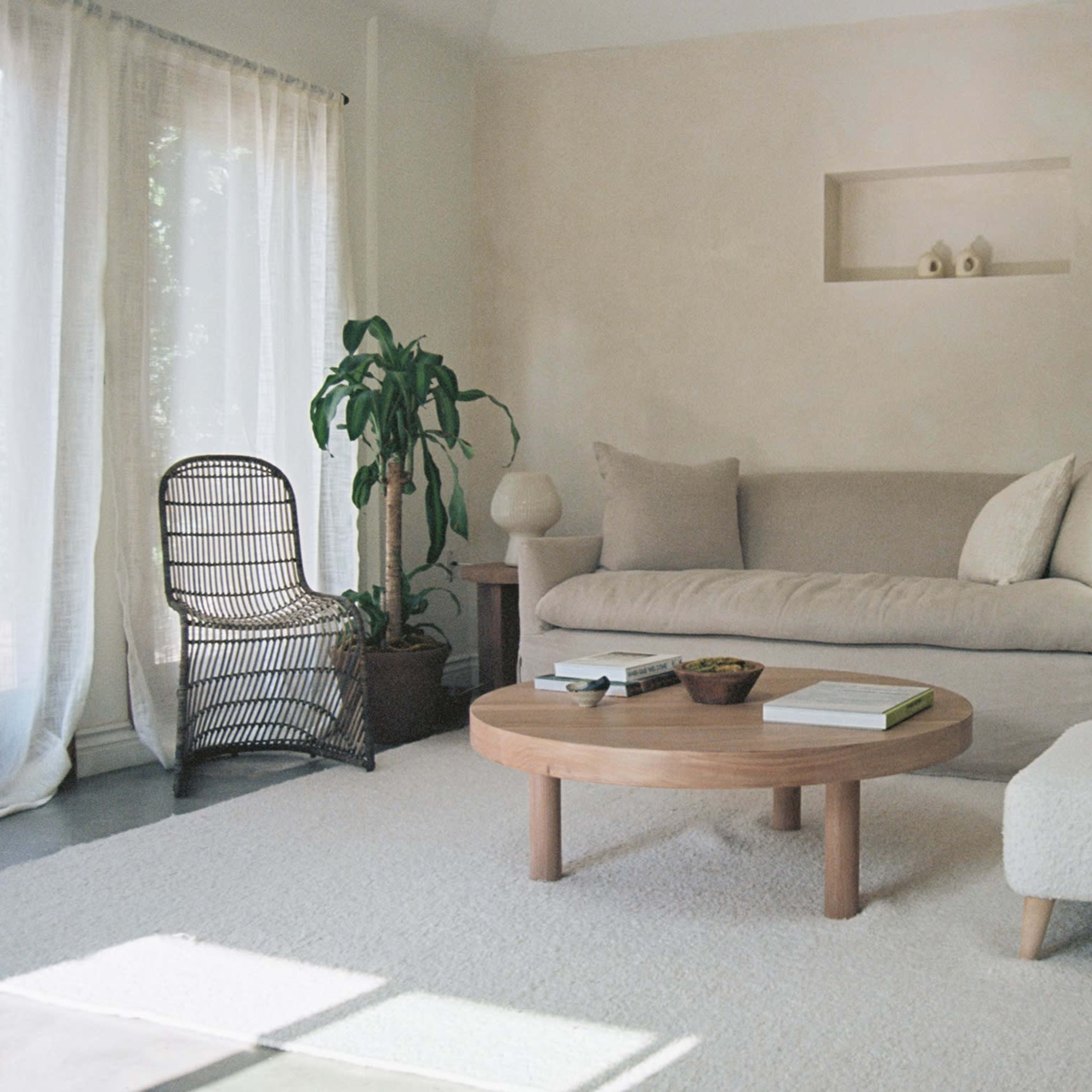 A light-colored living room features a simple sofa, a wooden coffee table, and a small ottoman, with a large plant and decorative elements.