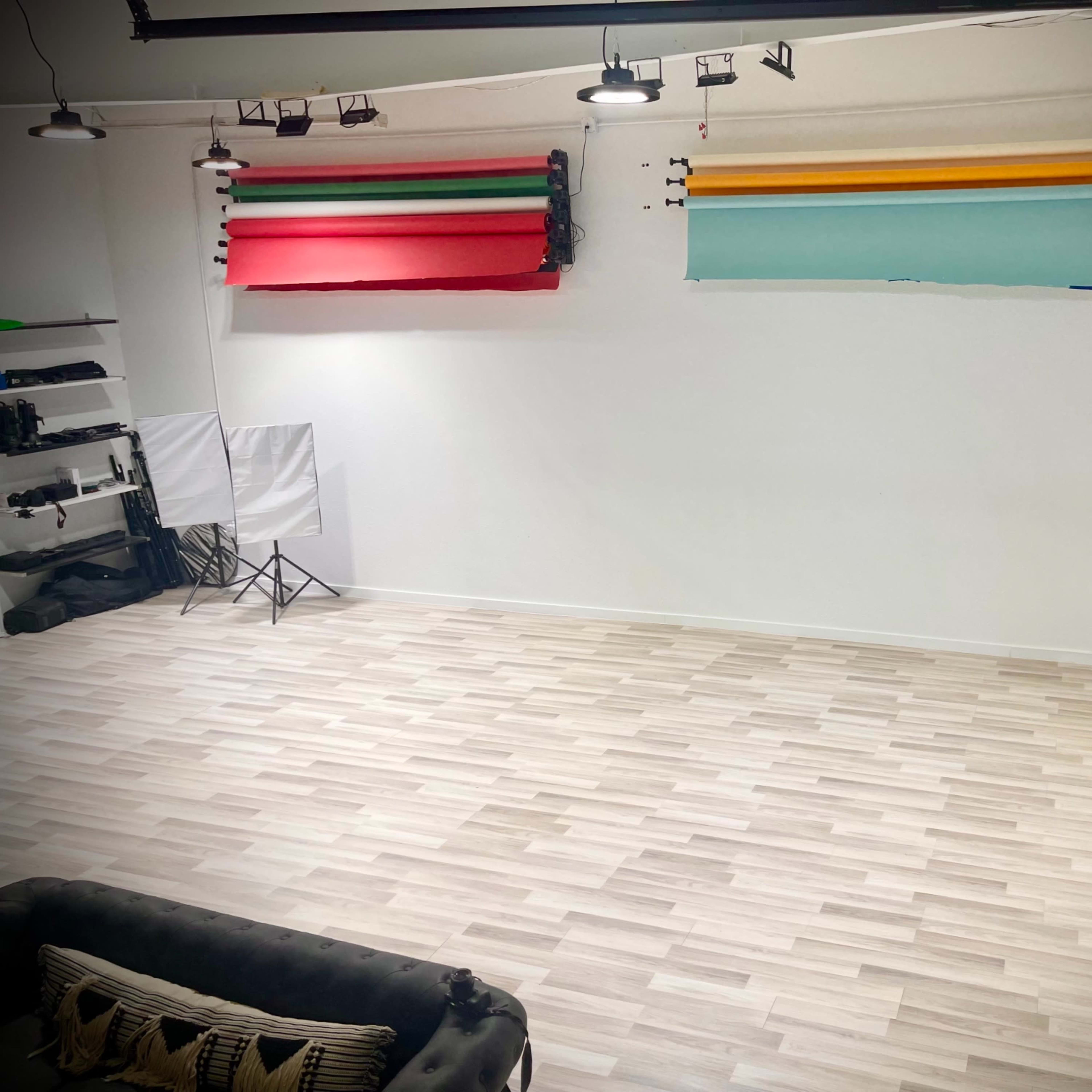A minimalist photography studio with a wooden floor, blank white walls, and colored backdrops hanging on the wall.