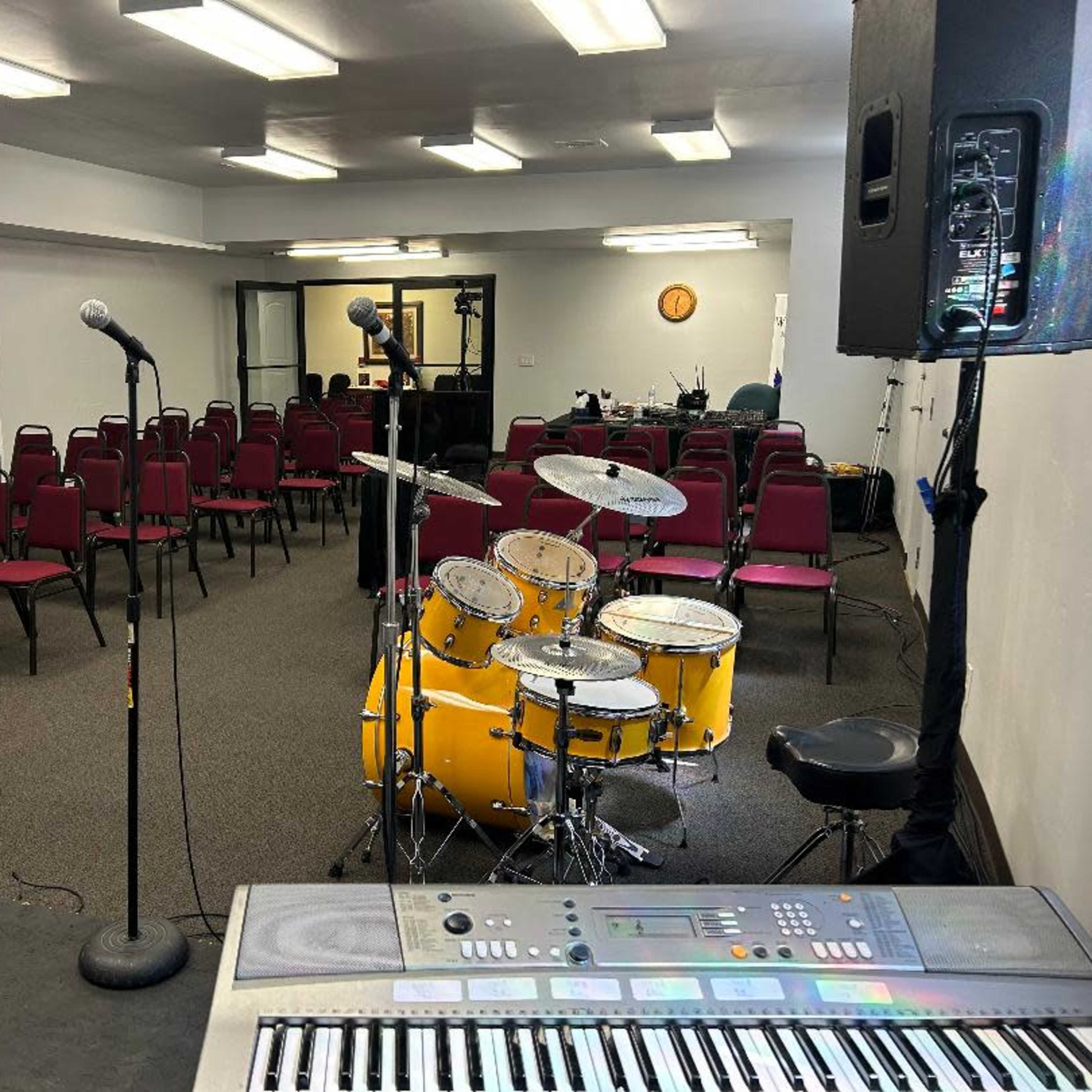 A setup for a performance or meeting, featuring a stage with a keyboard, a drum set, microphones, and rows of empty chairs arranged in a room.