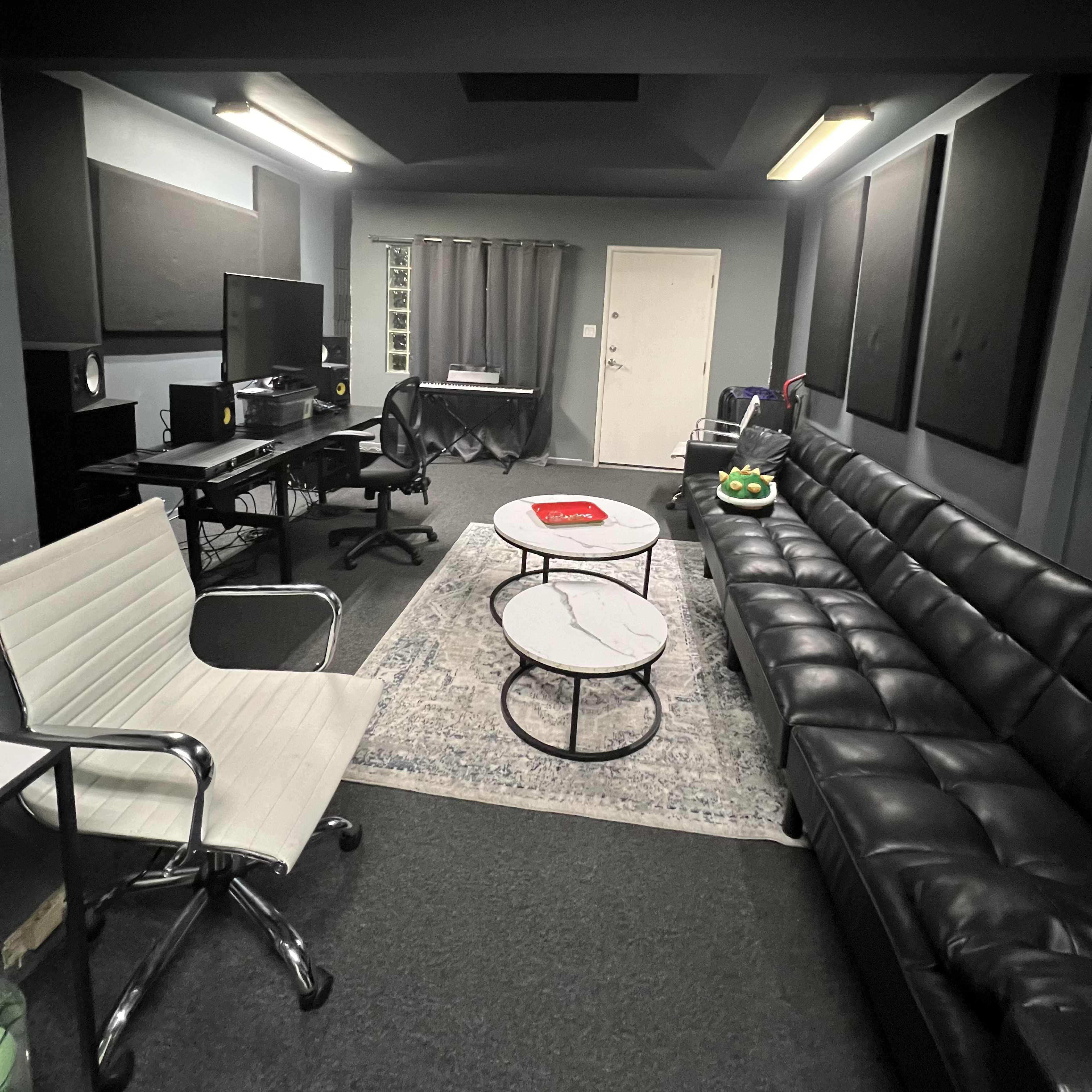 A small, modern office or studio space featuring a black leather couch, a couple of round tables, a desk with a computer, and soundproofing panels on the walls.