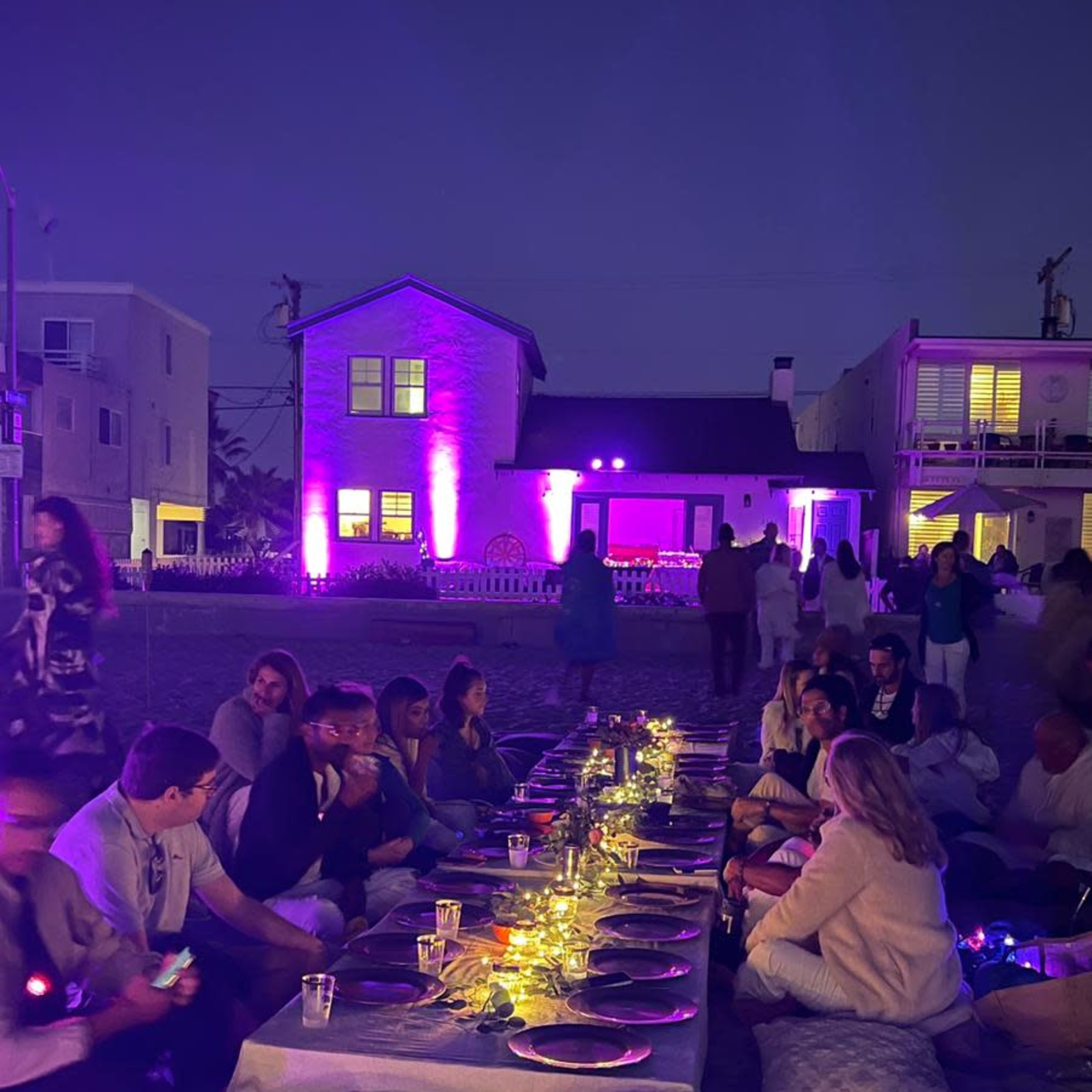 A group of people is seated around a long table on the ground, illuminated by purple lighting, in a nighttime outdoor setting.