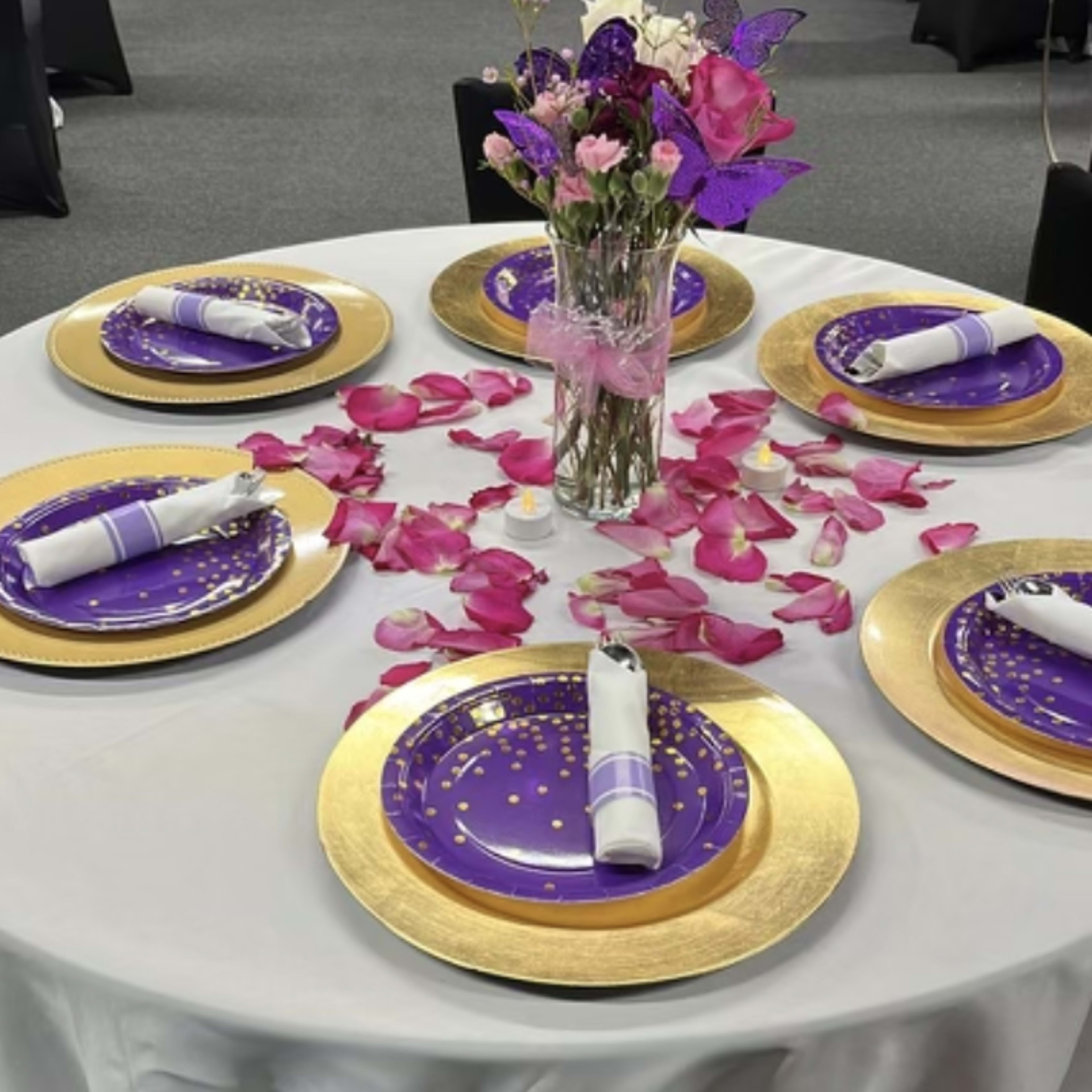 A round table is set with six purple plates on gold chargers, surrounded by pink rose petals and featuring a vase of flowers in the center.