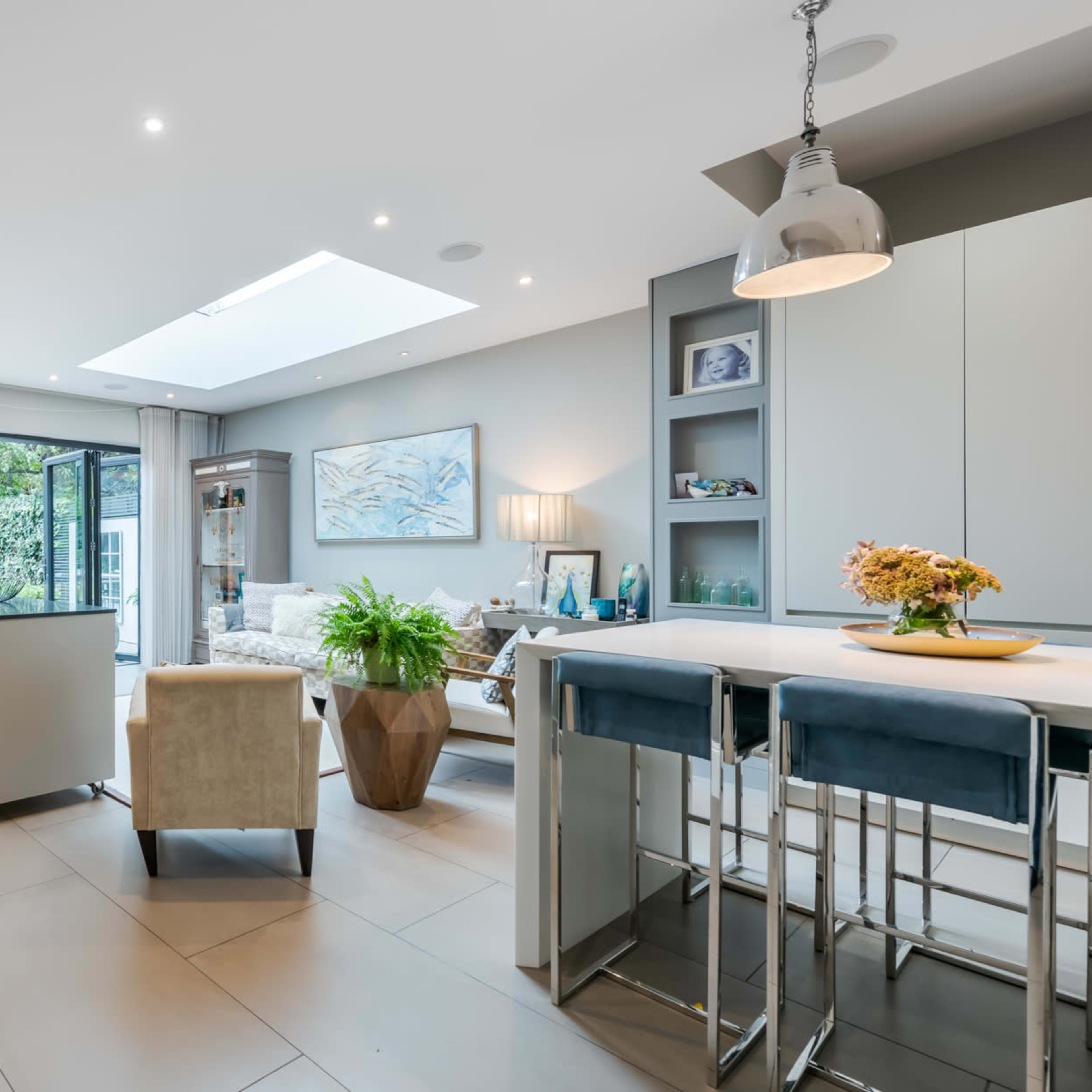 A spacious modern kitchen and living area with large glass doors opening to a garden, featuring elegant furniture and a neutral color scheme.