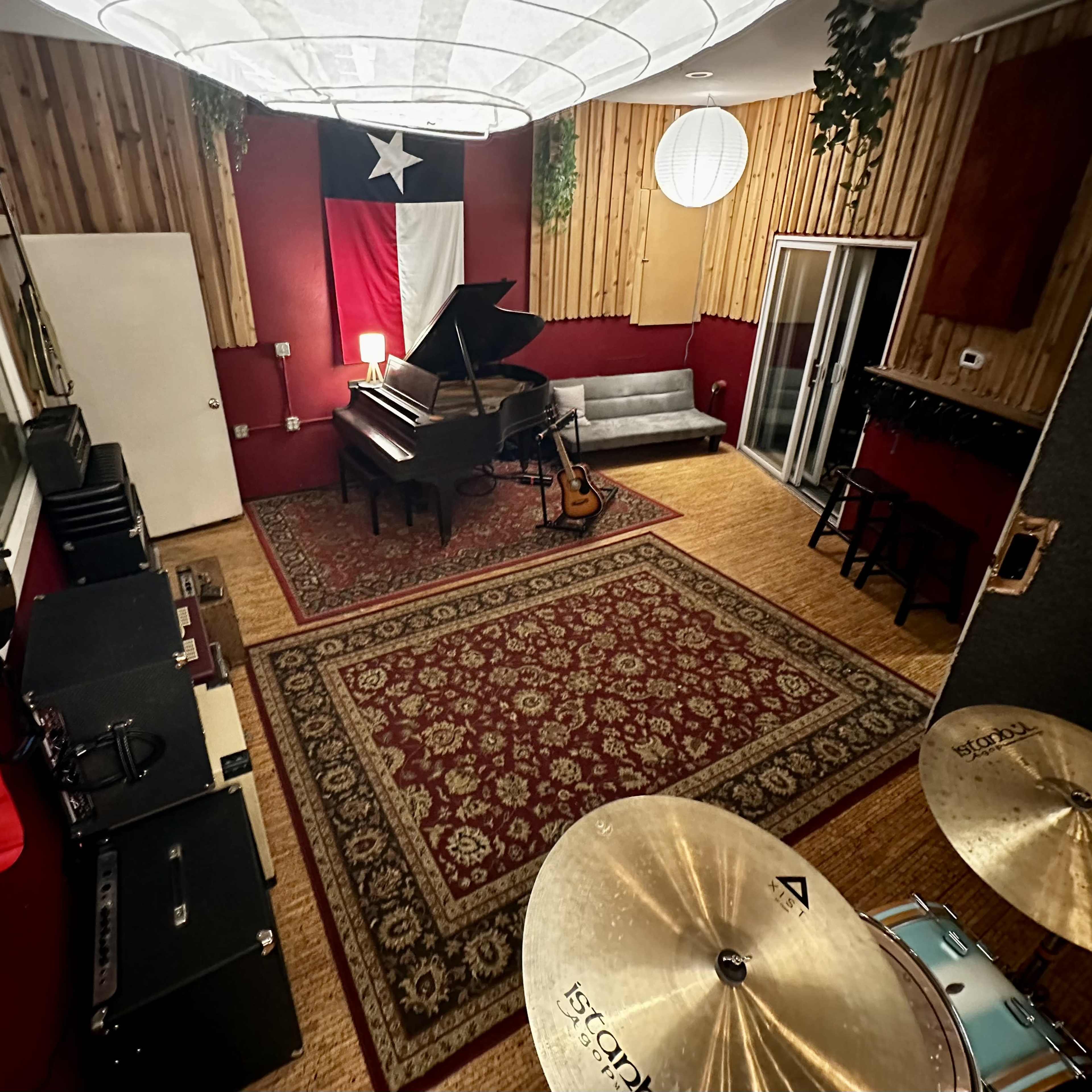 A music studio featuring a grand piano, a drum set, and various guitars alongside a patterned area rug in a room adorned with wooden paneling and a large Texas flag.