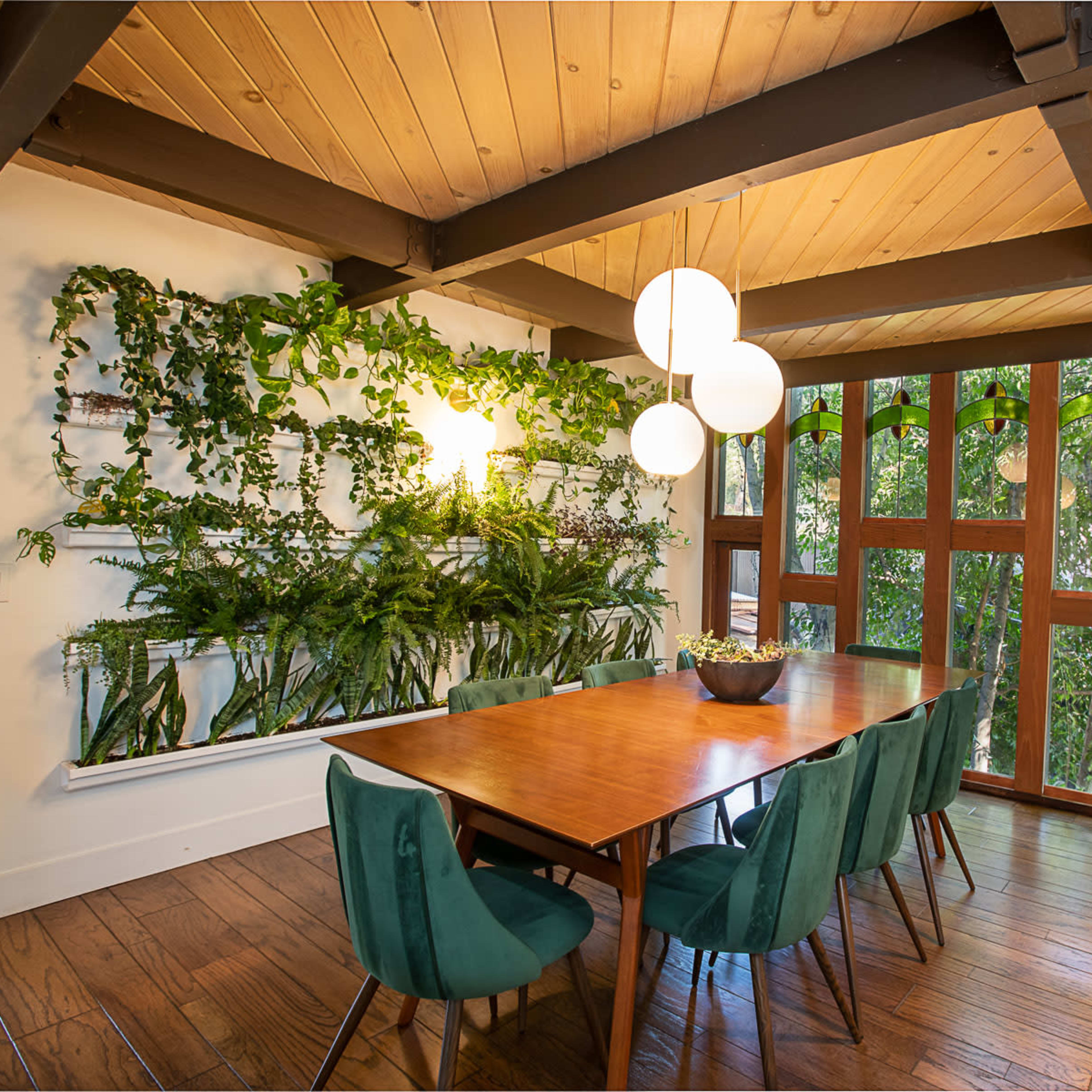A dining room features a long wooden table surrounded by green chairs, with a lush vertical garden on one wall and large windows allowing natural light.
