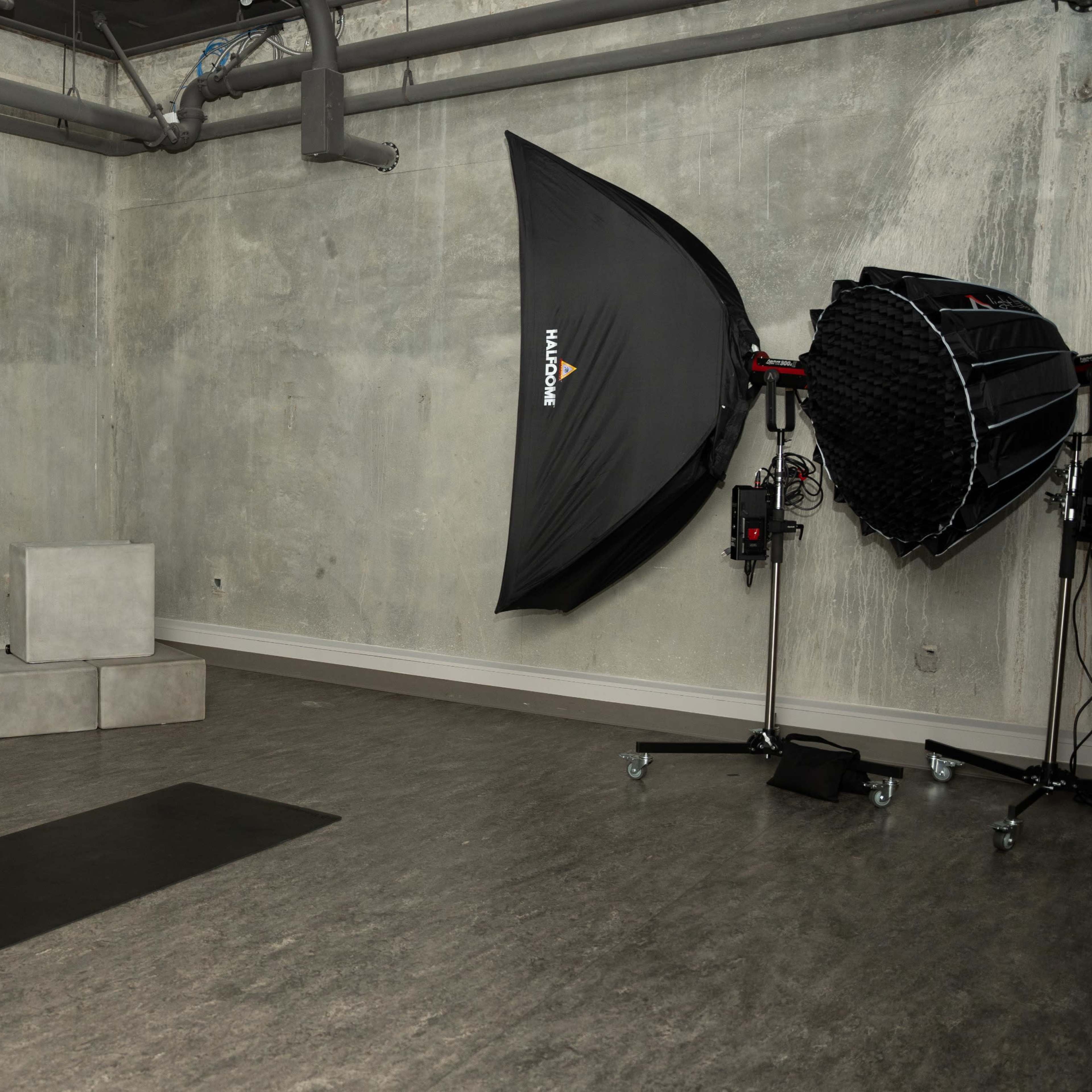 A minimalist studio space features a gray concrete wall, two concrete blocks, a black yoga mat on the floor, and a large softbox lighting setup in the corner.