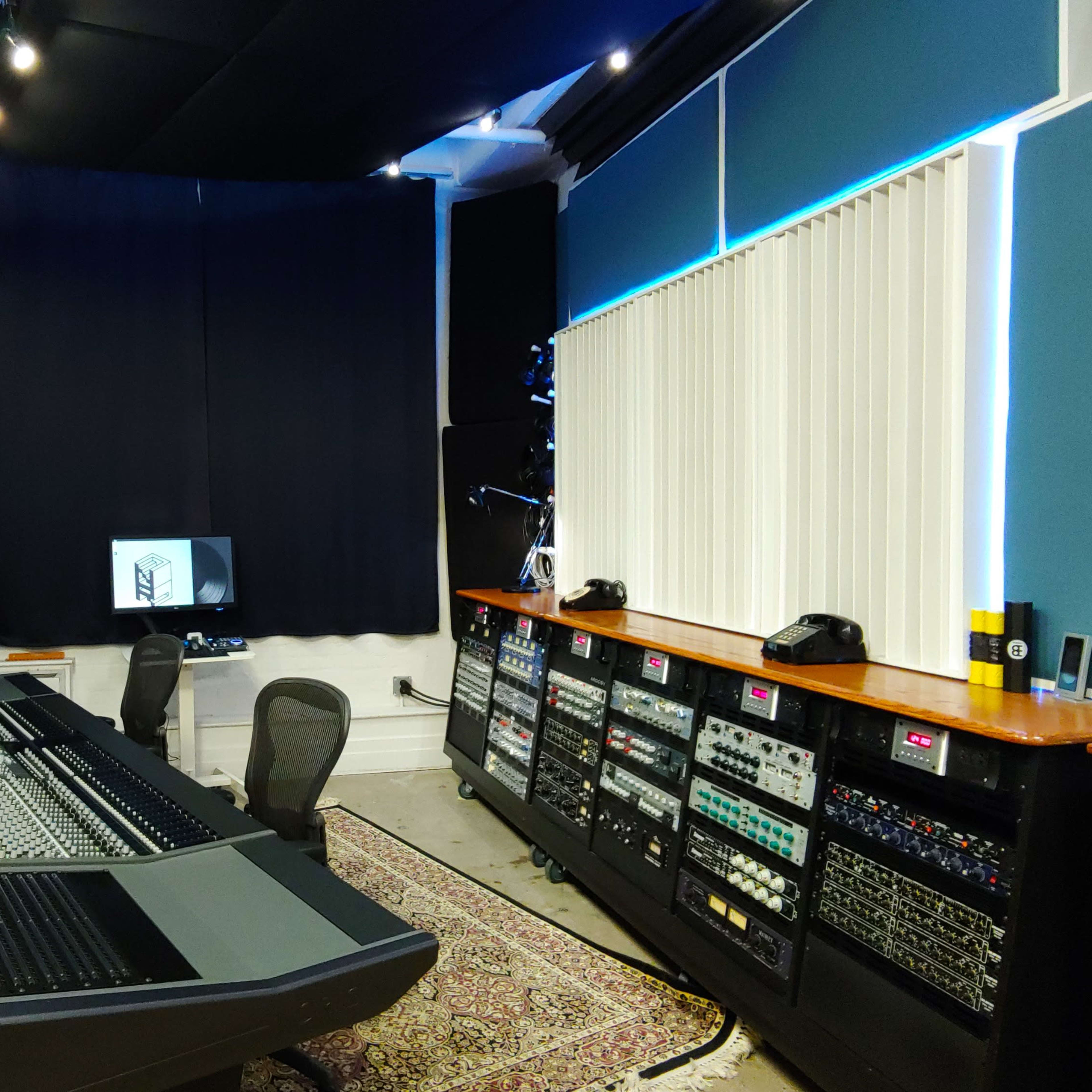 A modern recording studio features a large mixing console, various equipment racks, a computer display, and sound-proofed walls.