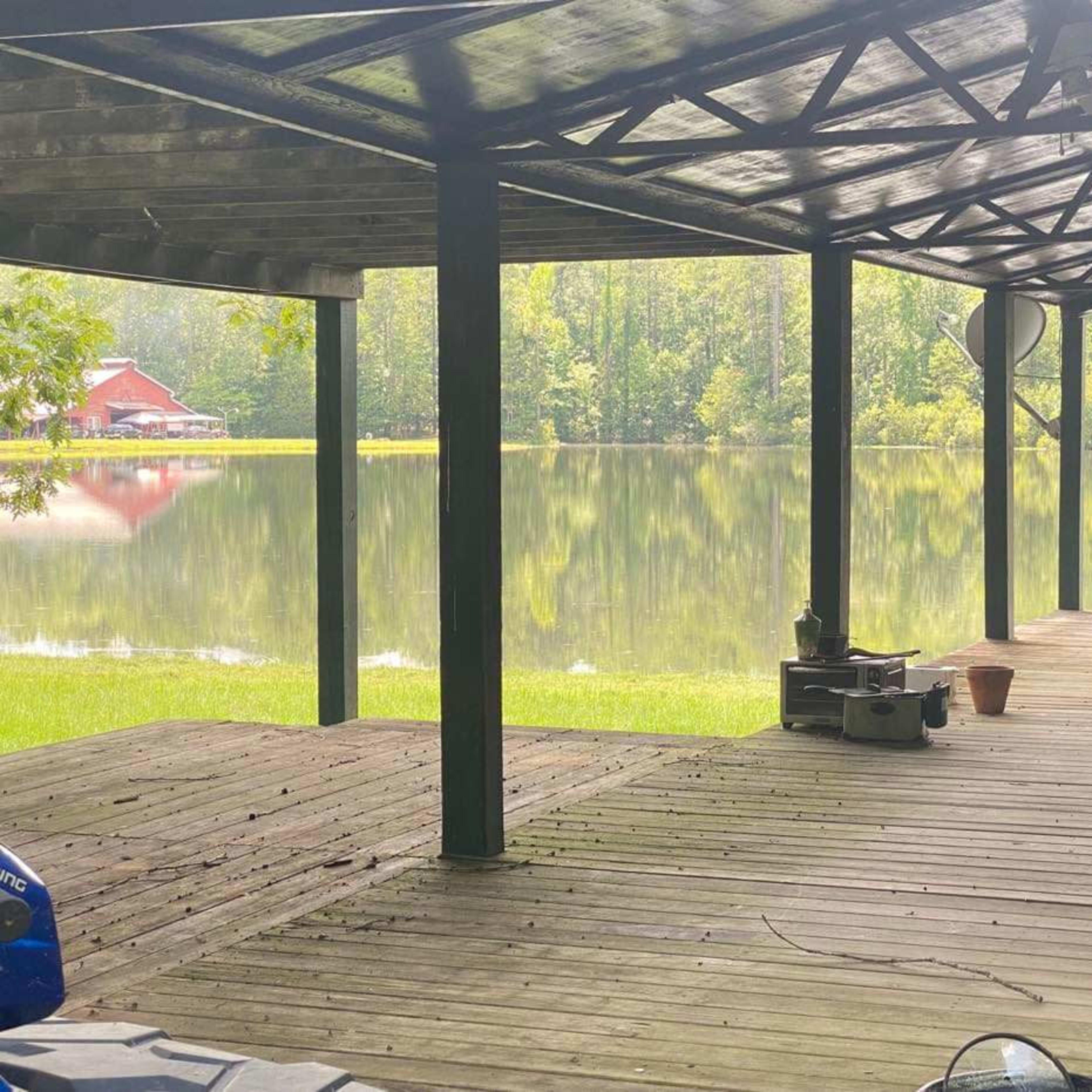 A wooden deck under a roof, overlooking a calm lake surrounded by trees, with a red building visible in the background.