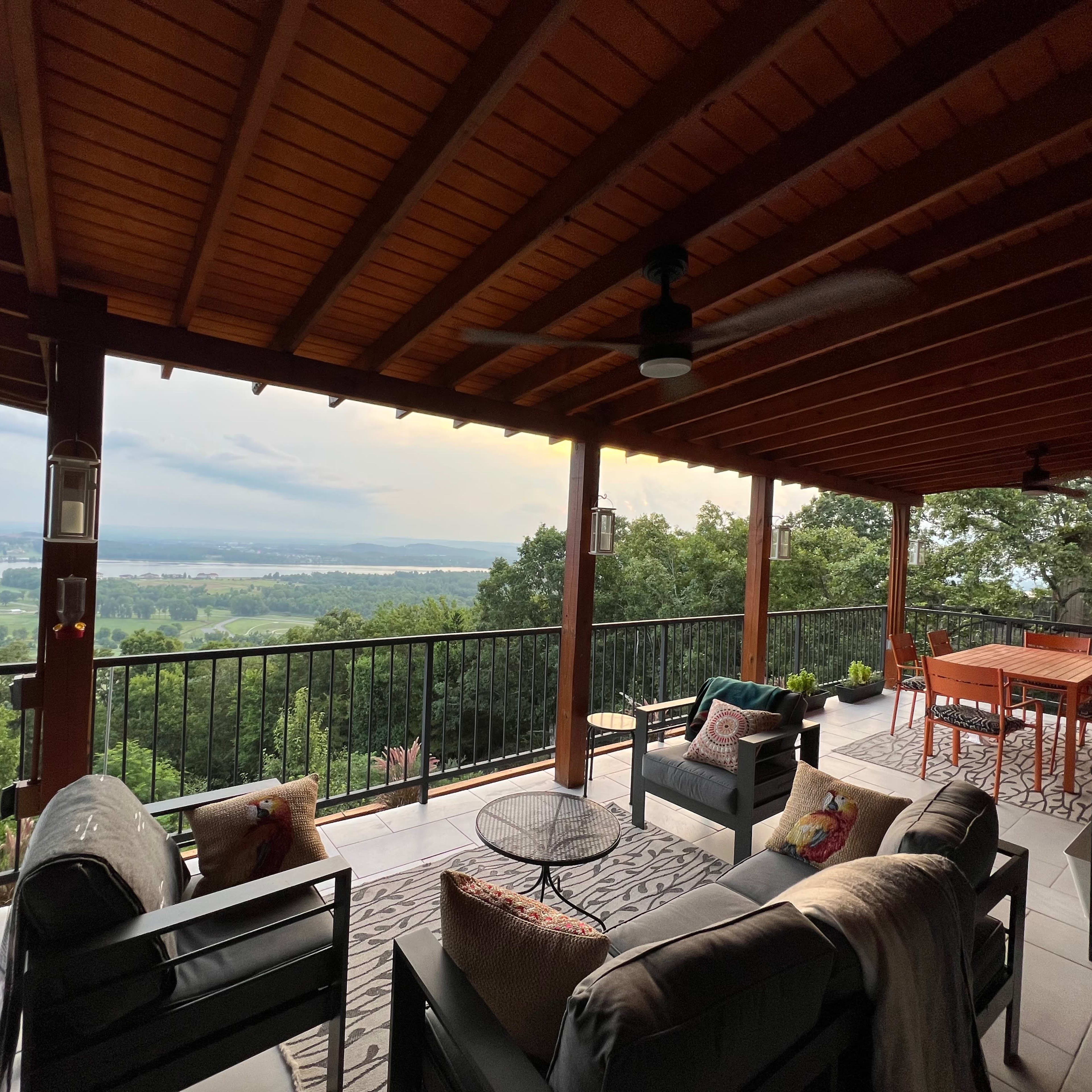 A covered patio with seating and a table, overlooking a green landscape under a cloudy sky.