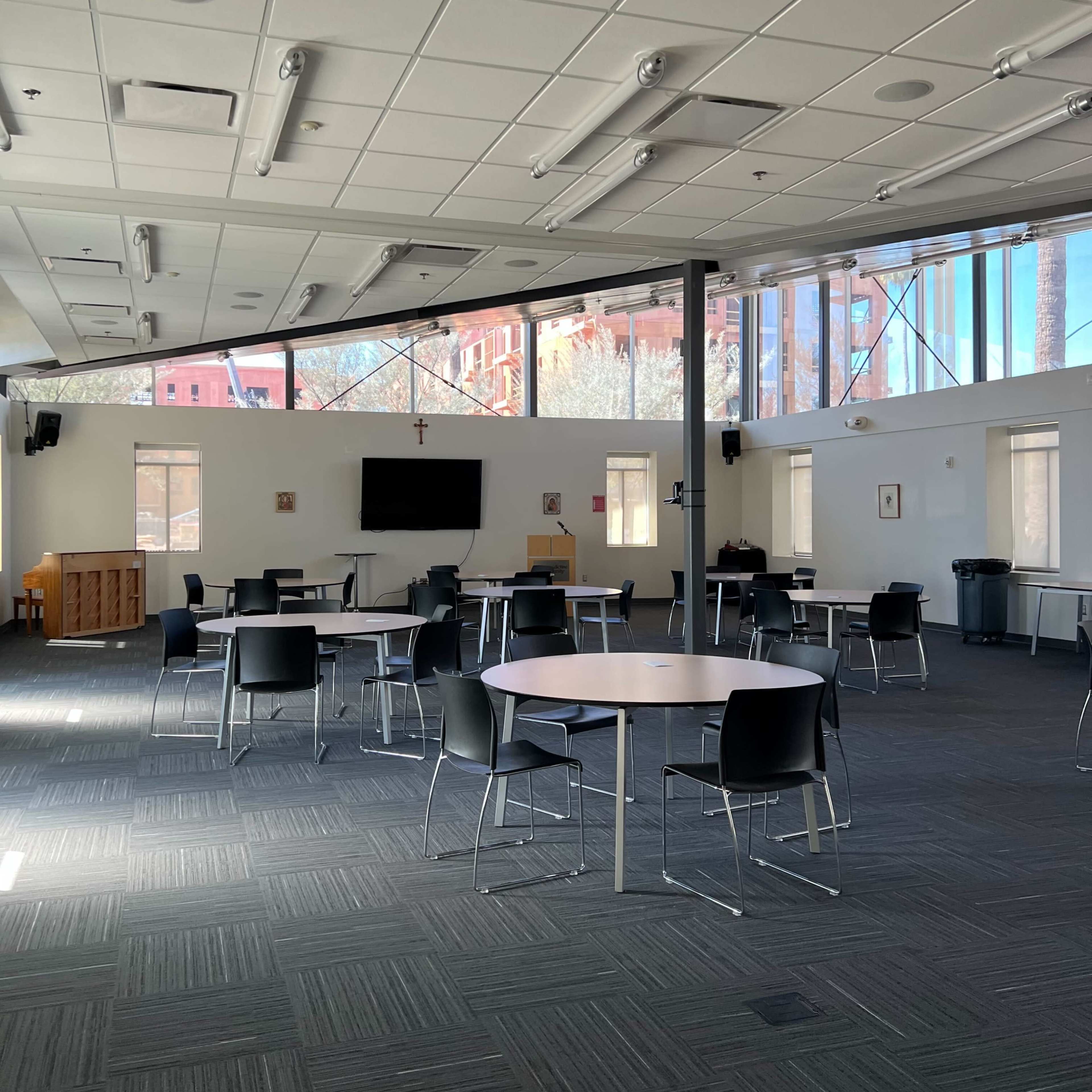 A spacious room with several round tables and black chairs arranged on a carpeted floor, illuminated by natural light from large windows.