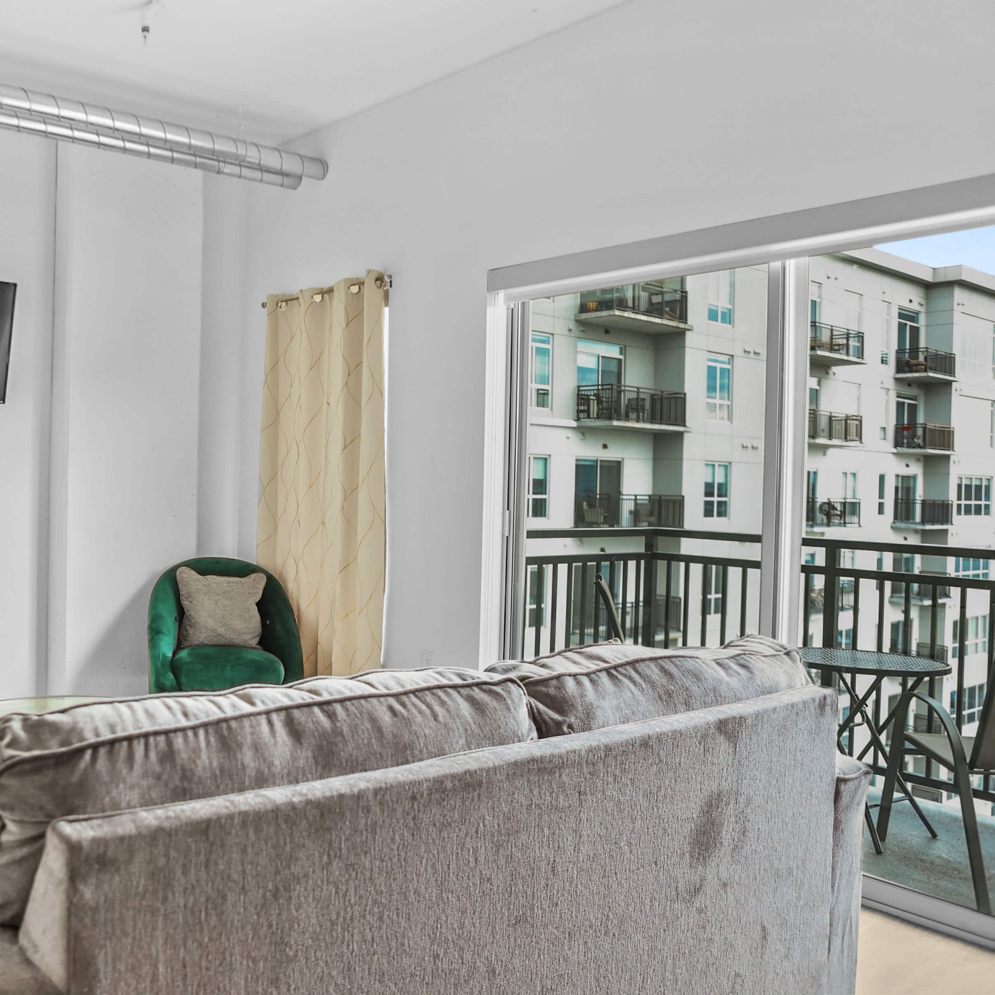A modern living room with two green chairs, a gray sofa, a wall-mounted television, and large windows overlooking a balcony.