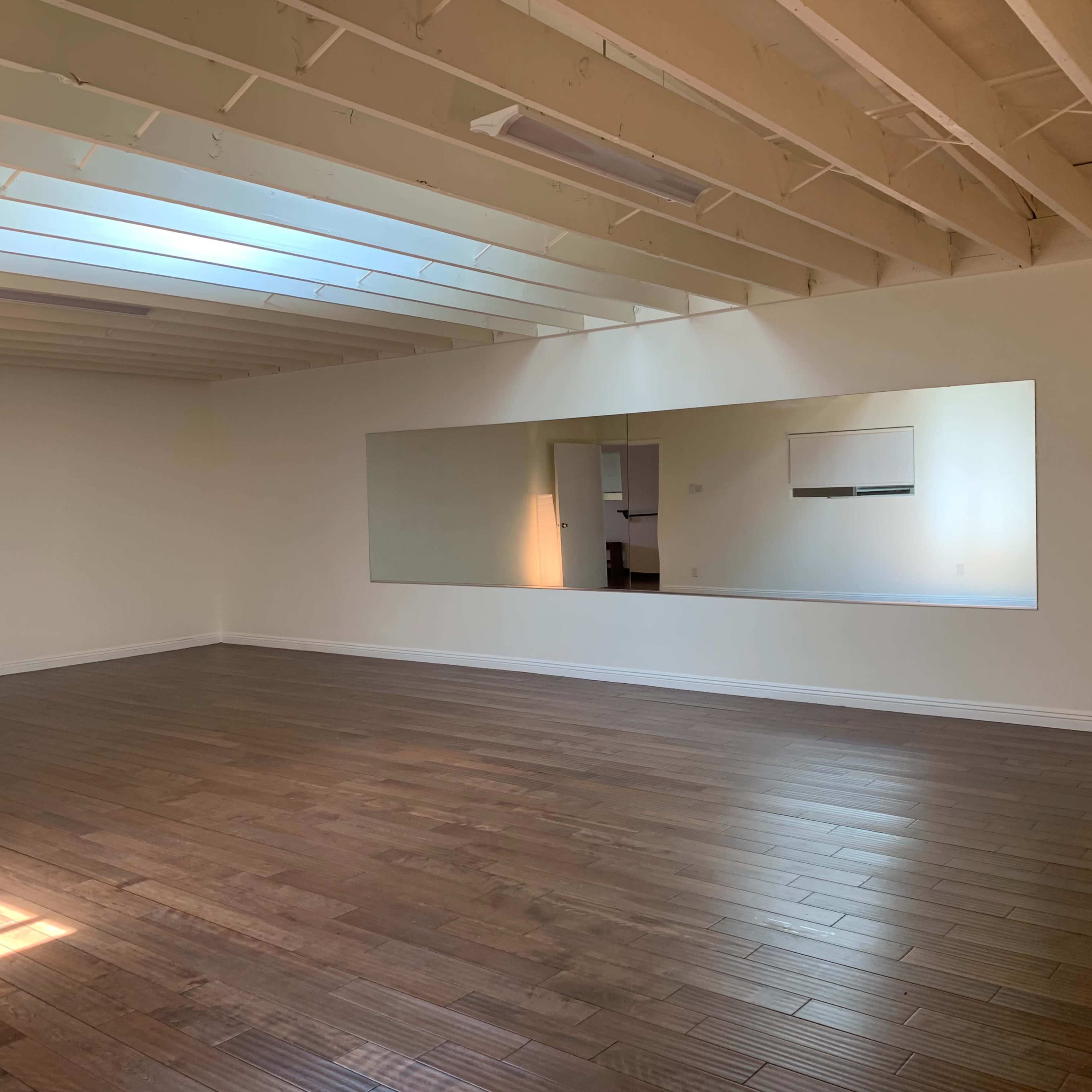 A spacious room with wooden floors, a large mirror on one wall, and natural light coming through a skylight.