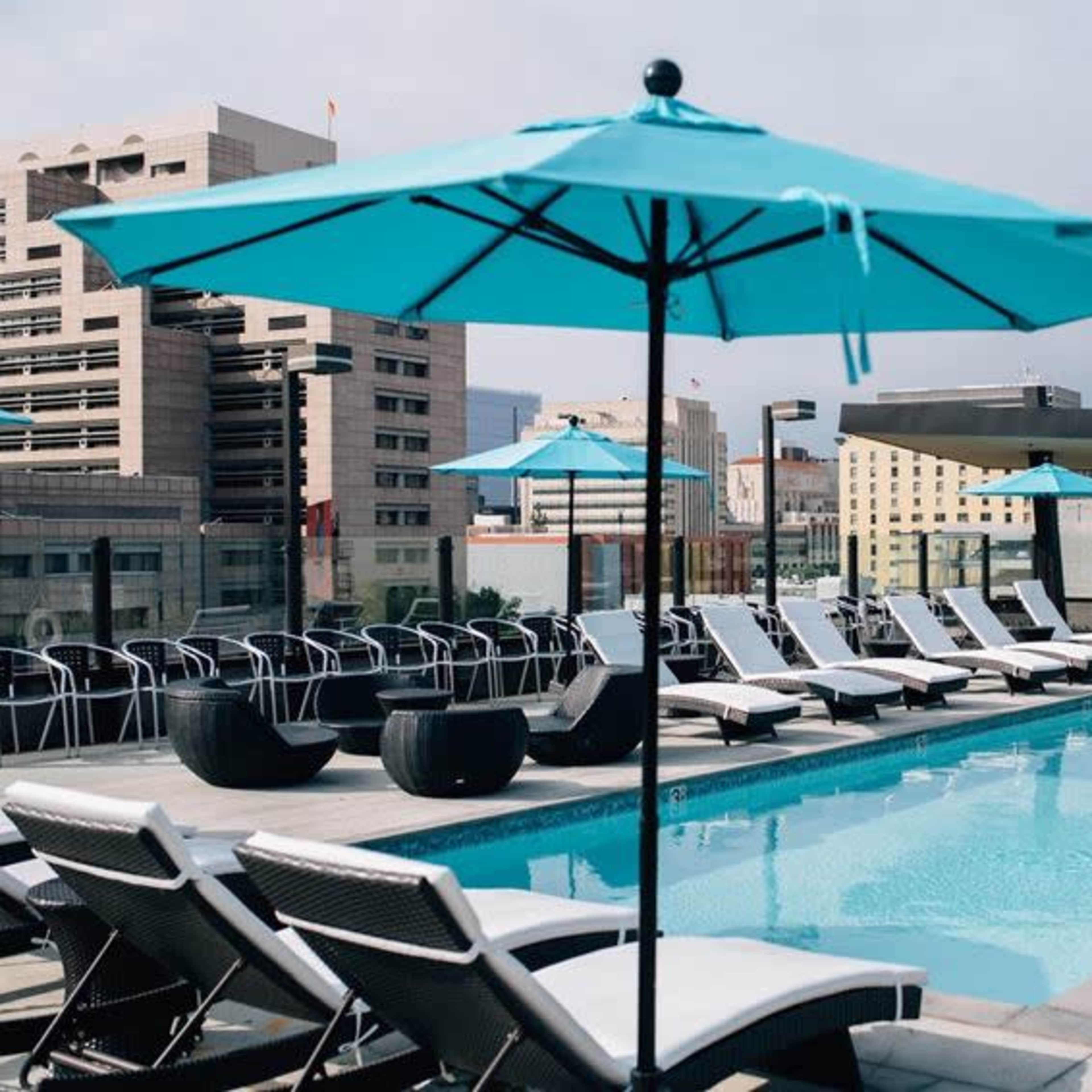 A rooftop pool area features lounge chairs and turquoise umbrellas against a backdrop of city buildings.