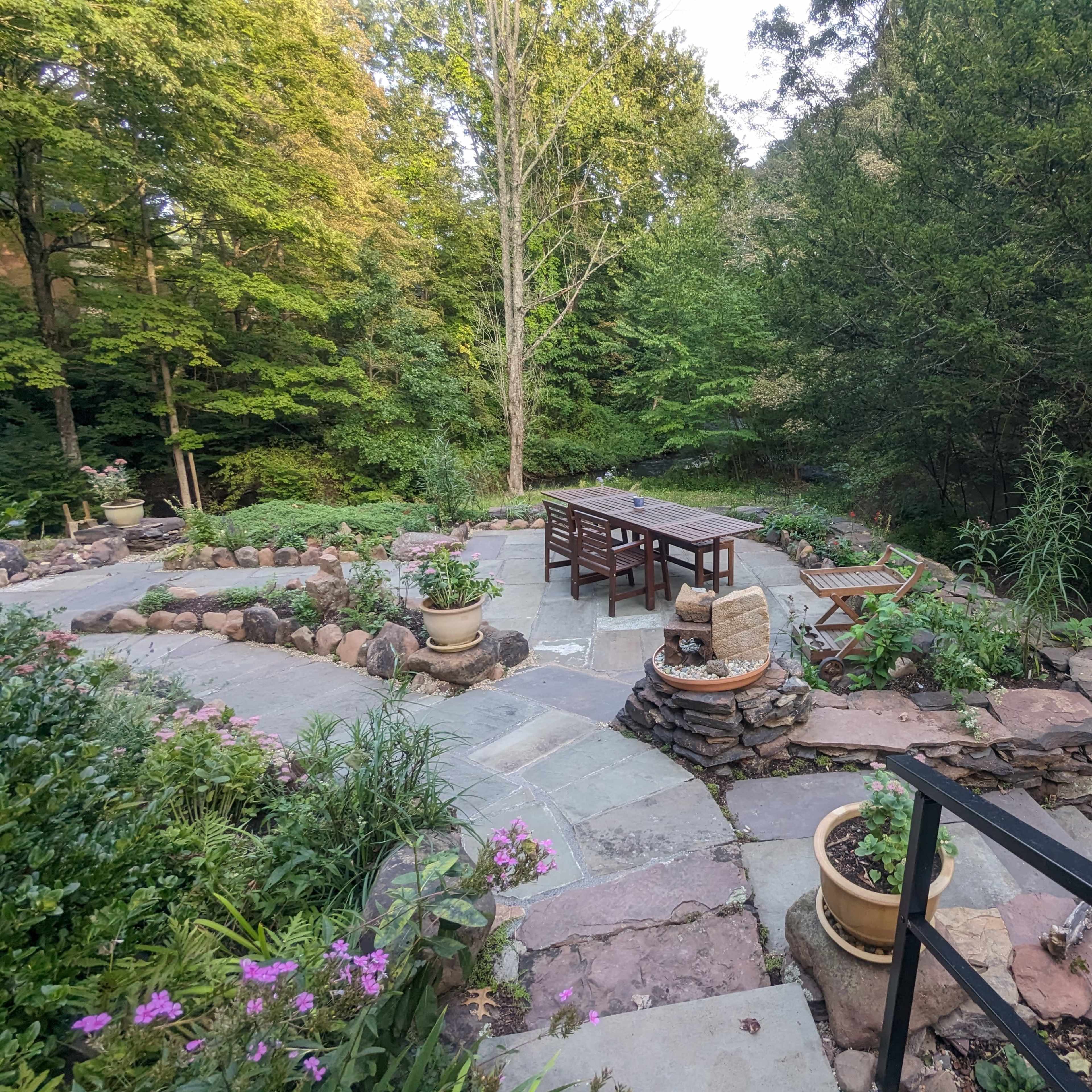 A landscaped outdoor area with a stone patio, a wooden table and chairs, and surrounding greenery beside a stream.
