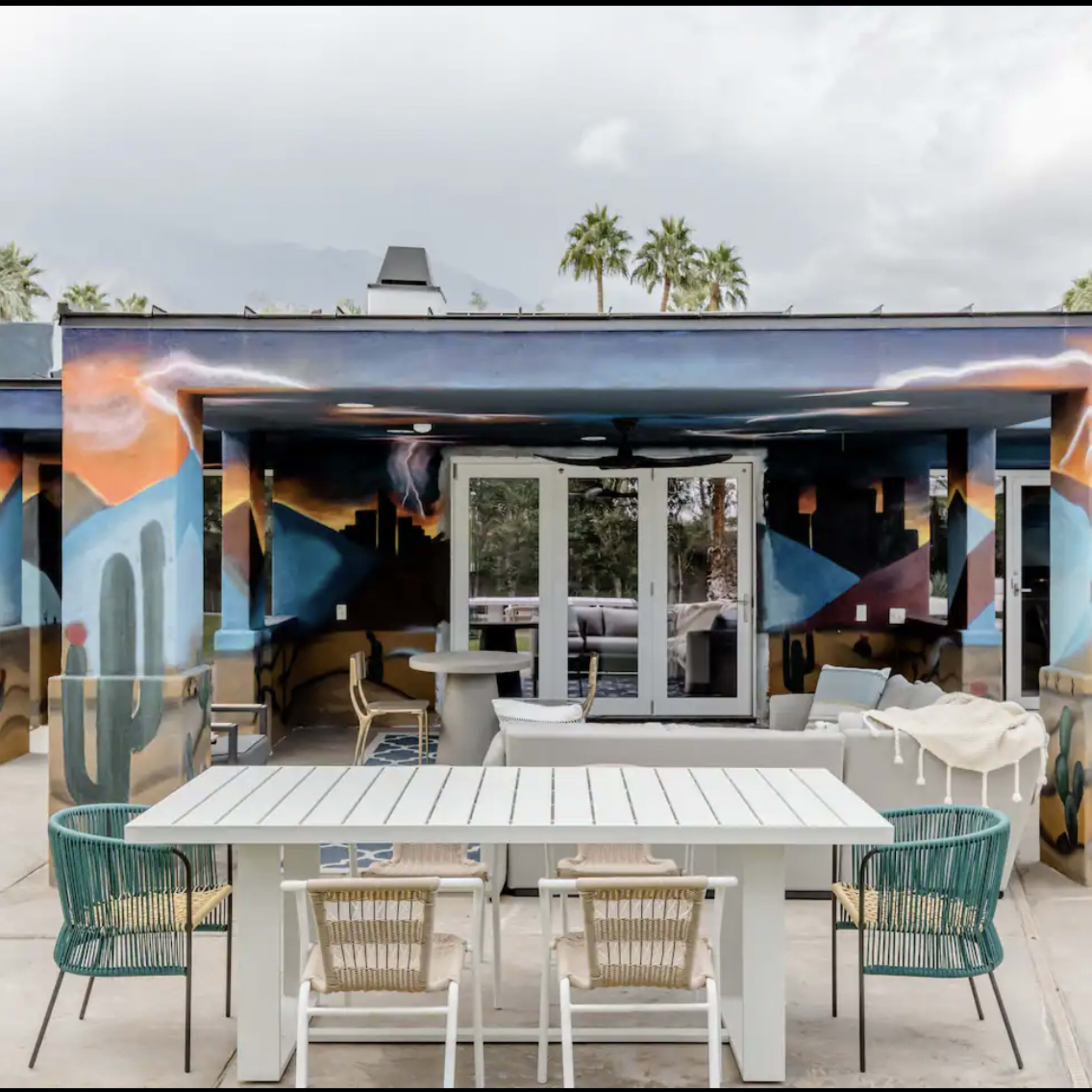 A modern outdoor space features a large table with chairs, surrounded by walls displaying vibrant murals and palm trees in the background.