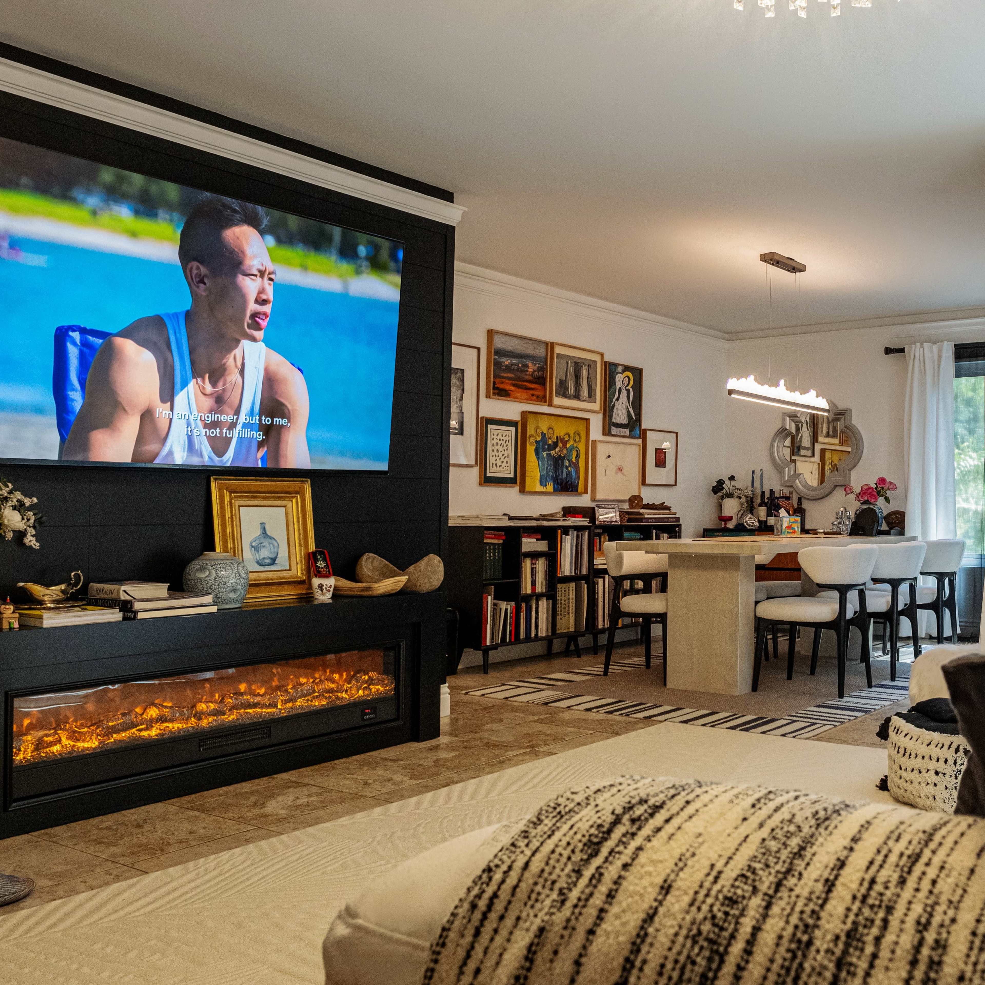 A modern living room featuring a large wall-mounted television displaying a scene, a dining area with a table and chairs, and a fireplace beneath the TV.