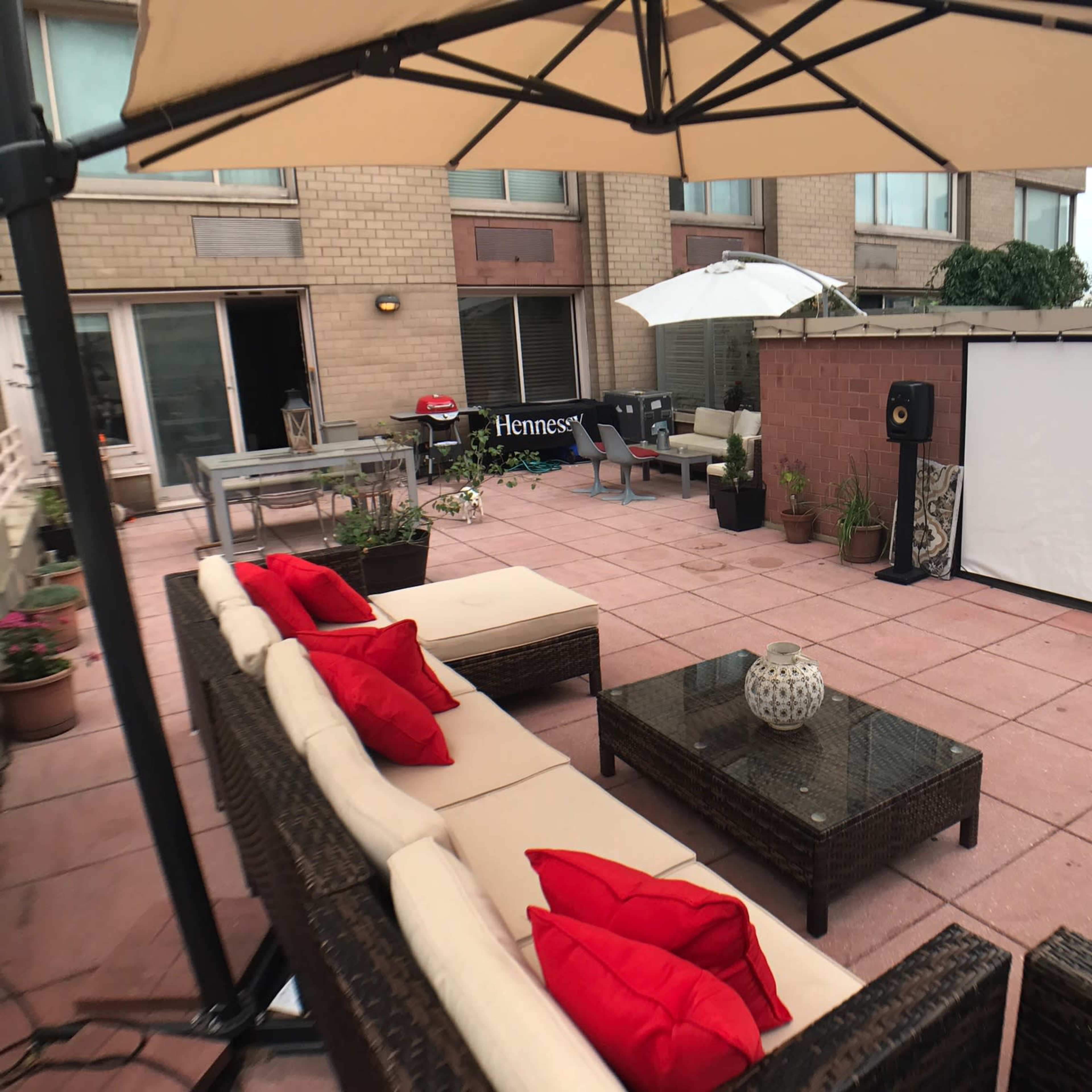 A patio area with a sectional sofa, coffee table, and a large white screen for projected viewing, surrounded by potted plants and an outdoor umbrella.