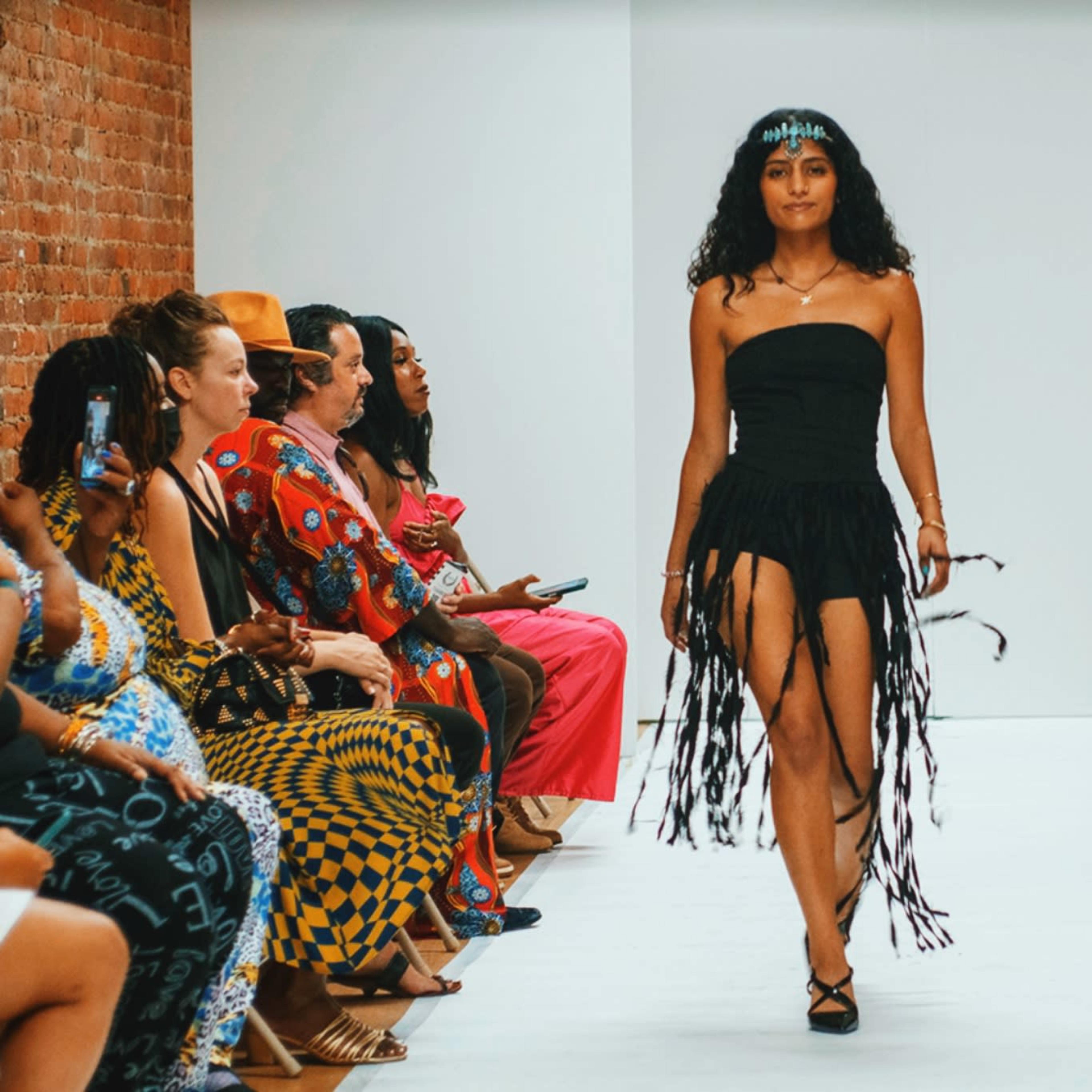 A model walks down a runway wearing a fringed black dress while an audience sits on either side.