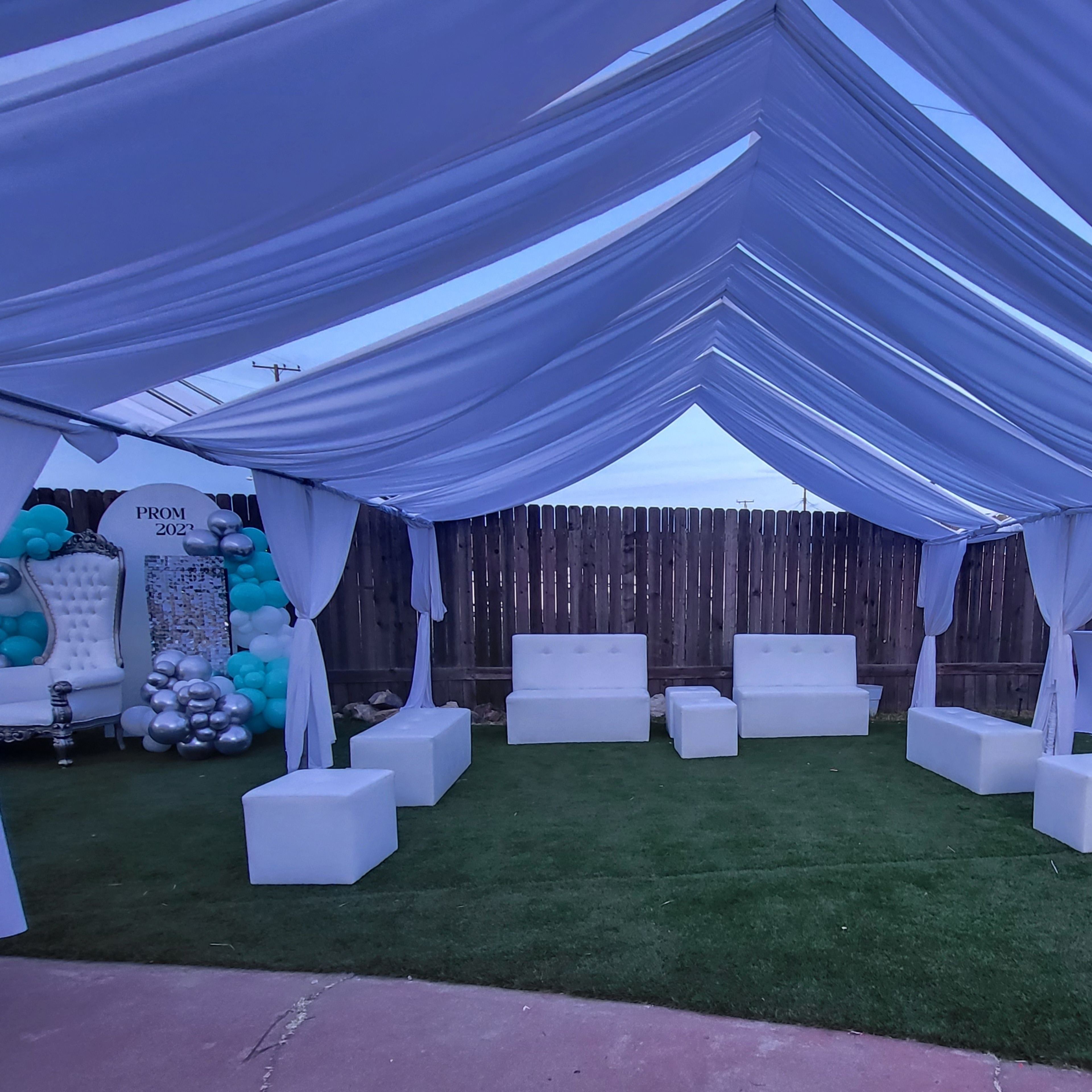 A beautifully arranged outdoor event space features white draped canopies, green grass, and white lounge seating with decorative elements in the background.