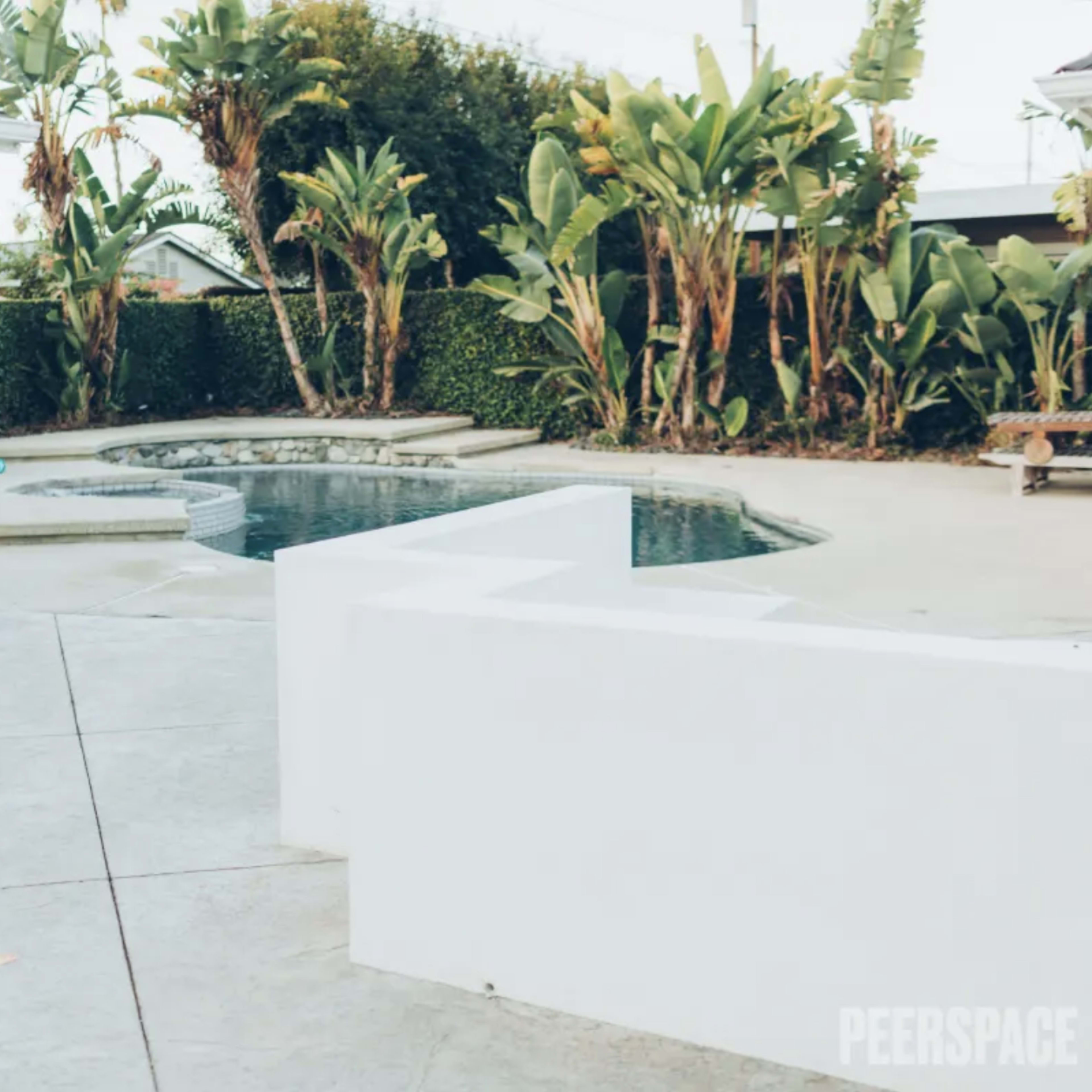 A swimming pool area enclosed by tropical plants and a white building with a pink door.