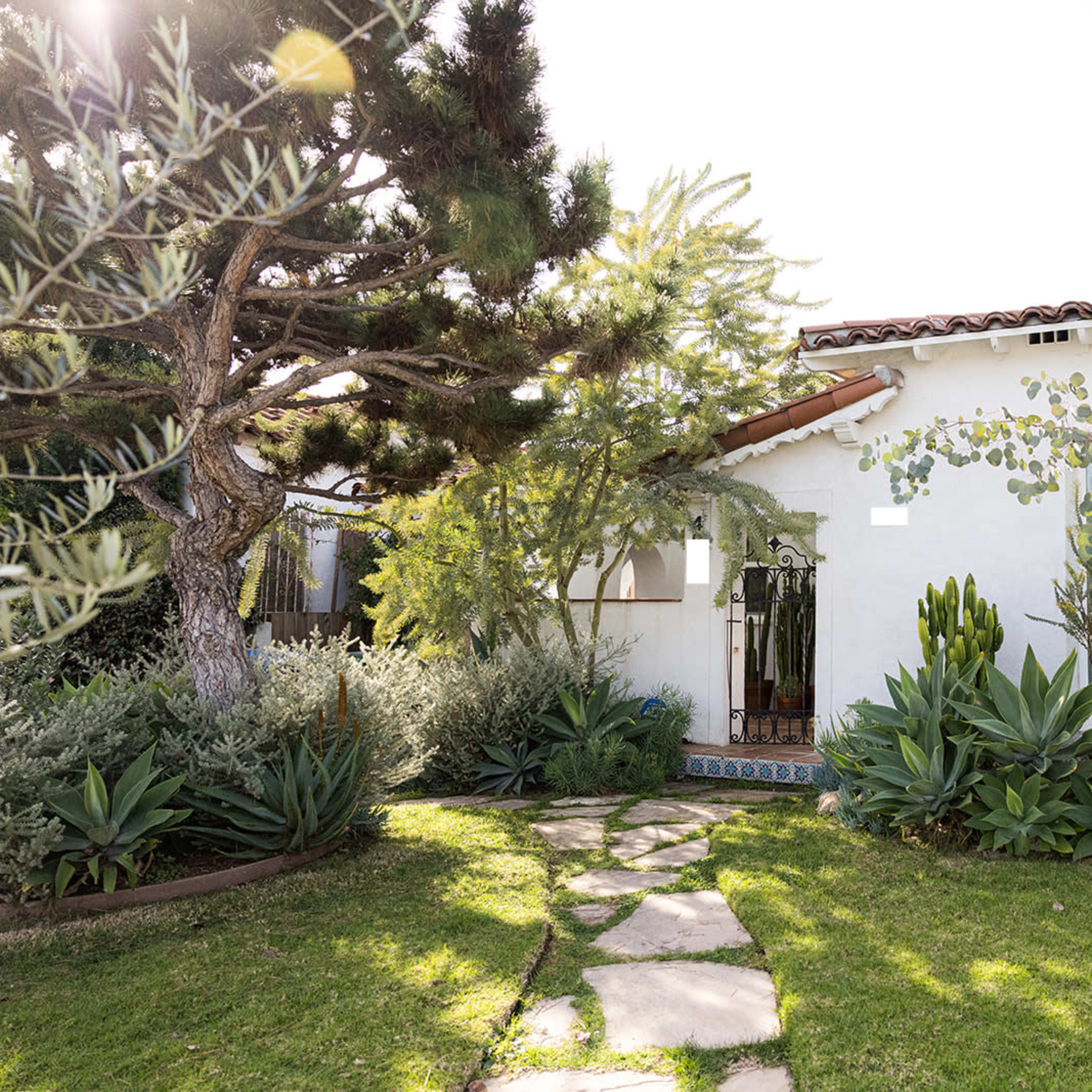 A well-maintained white house with a sloped roof is surrounded by lush greenery and a stone pathway leading to the entrance.