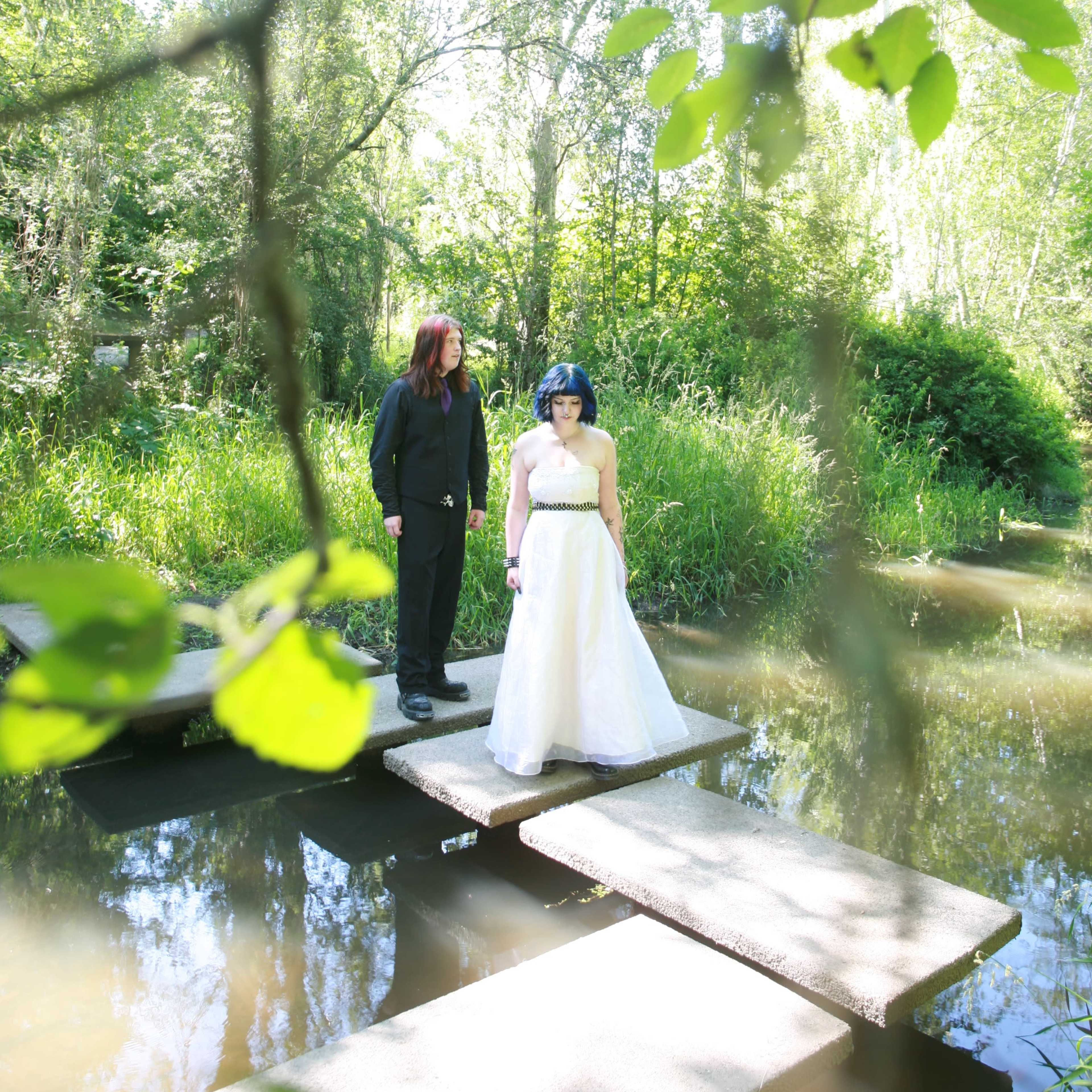 Two individuals stand on stepping stones across a calm body of water in a lush, green setting.