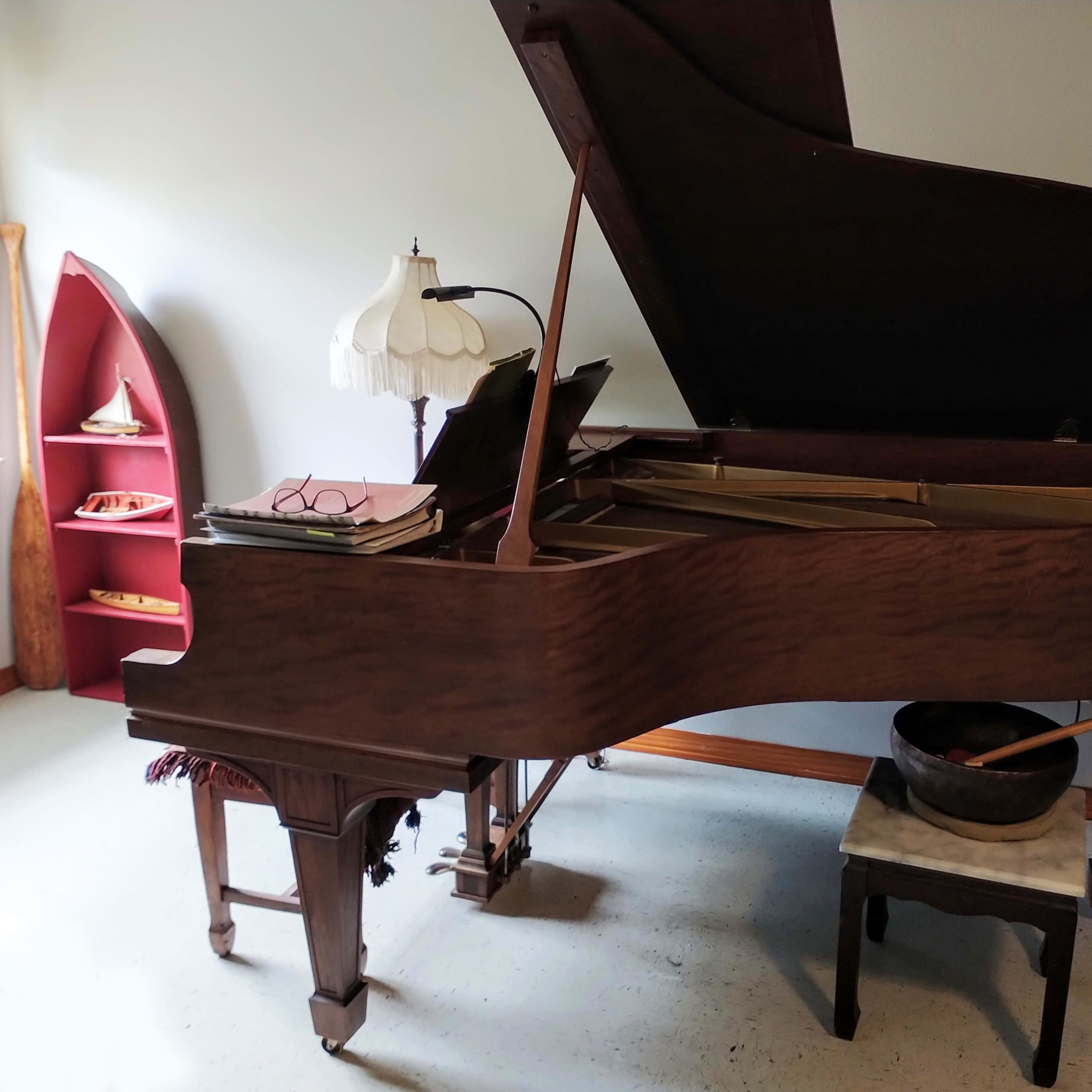A grand piano with an open lid sits beside a lamp and a small table in a room featuring wooden shelves.