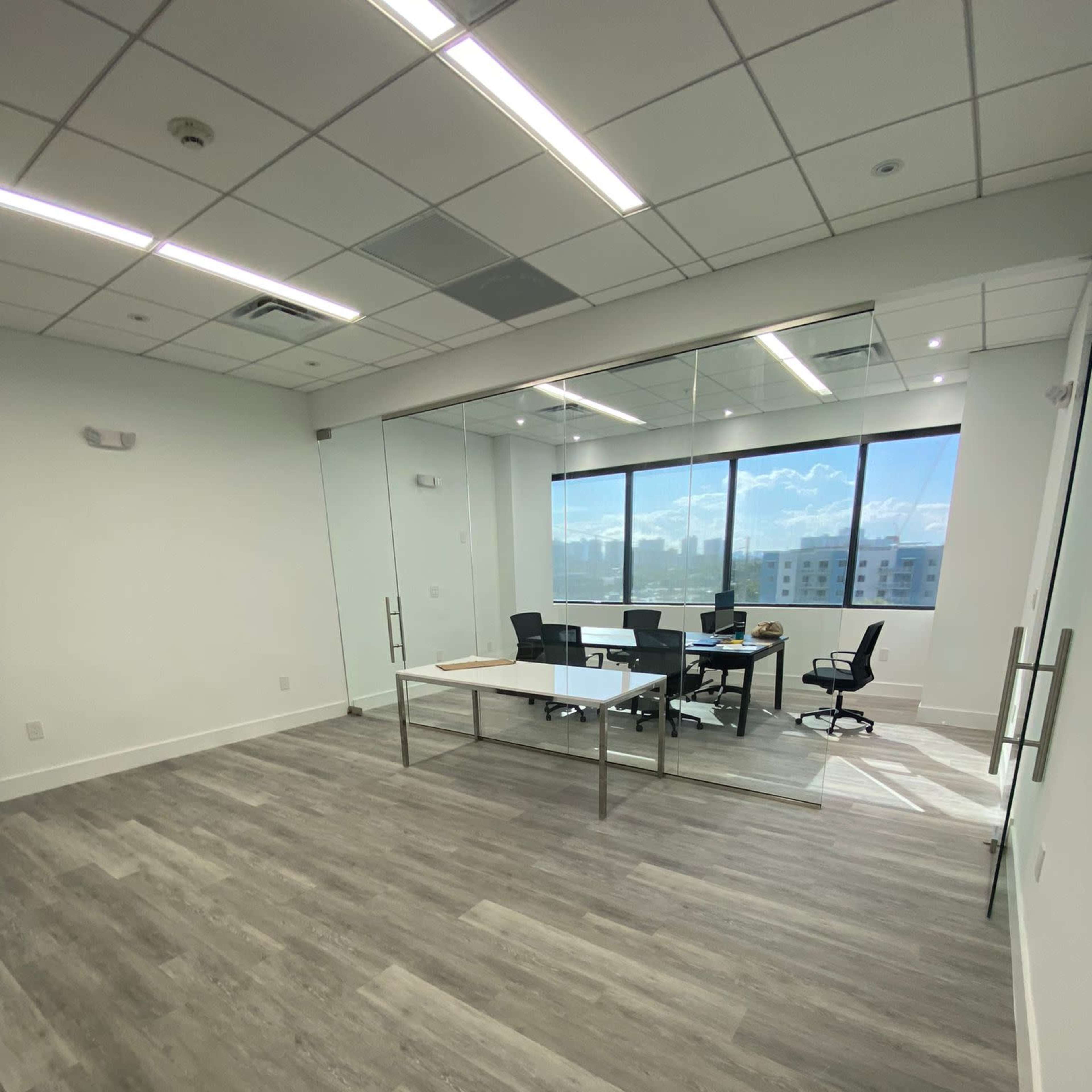 An empty modern office space features a glass-walled room with a desk, a computer, and two chairs, illuminated by natural light from large windows.