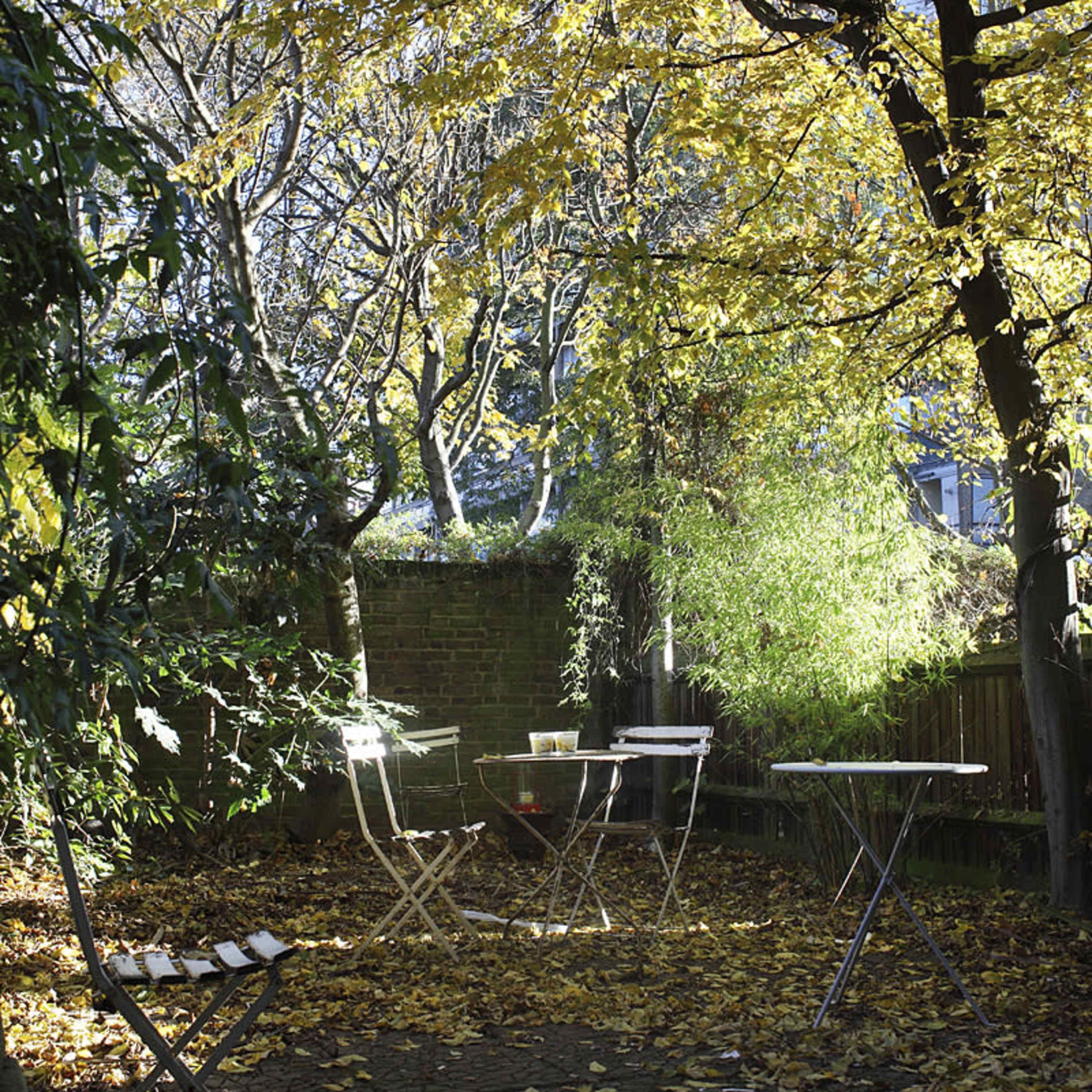 A small outdoor seating area is surrounded by trees with autumn leaves, featuring two metal tables and chairs on a stone pathway covered with fallen leaves.