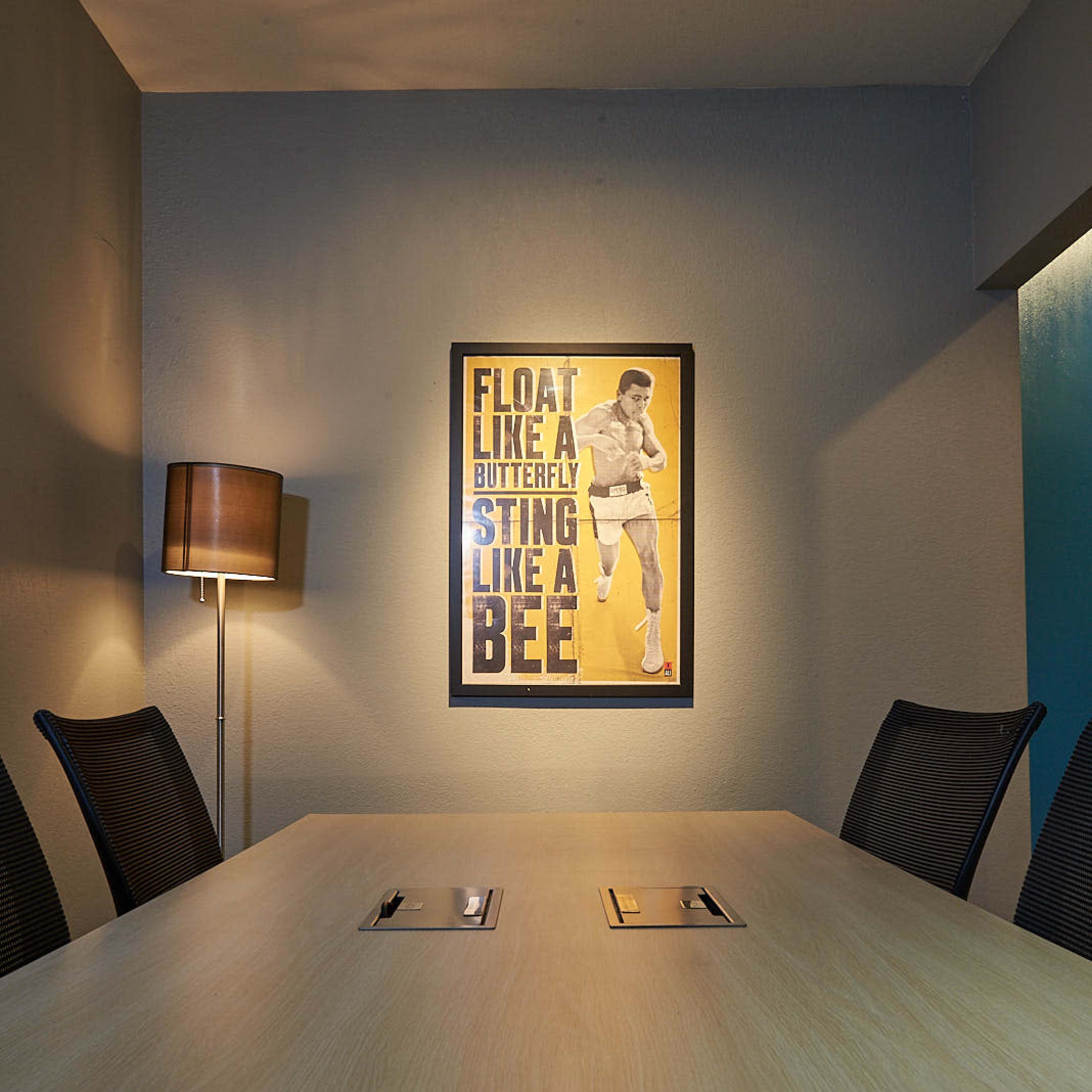 A meeting room features a wooden table surrounded by chairs, with a lamp and a motivational poster on the wall.
