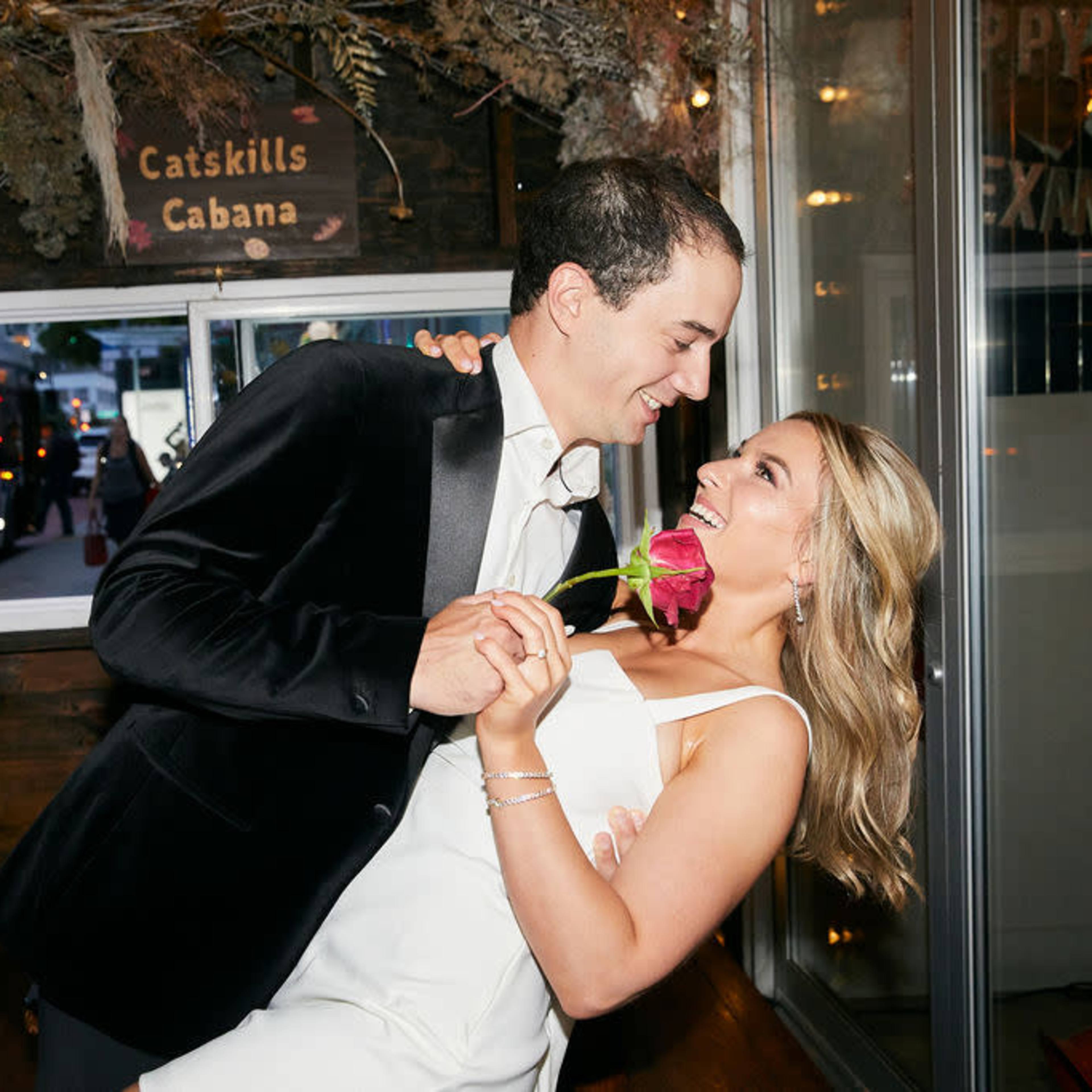 A man in a black suit and a woman in a white dress are dancing together inside an elegantly decorated venue, with the woman holding a rose.