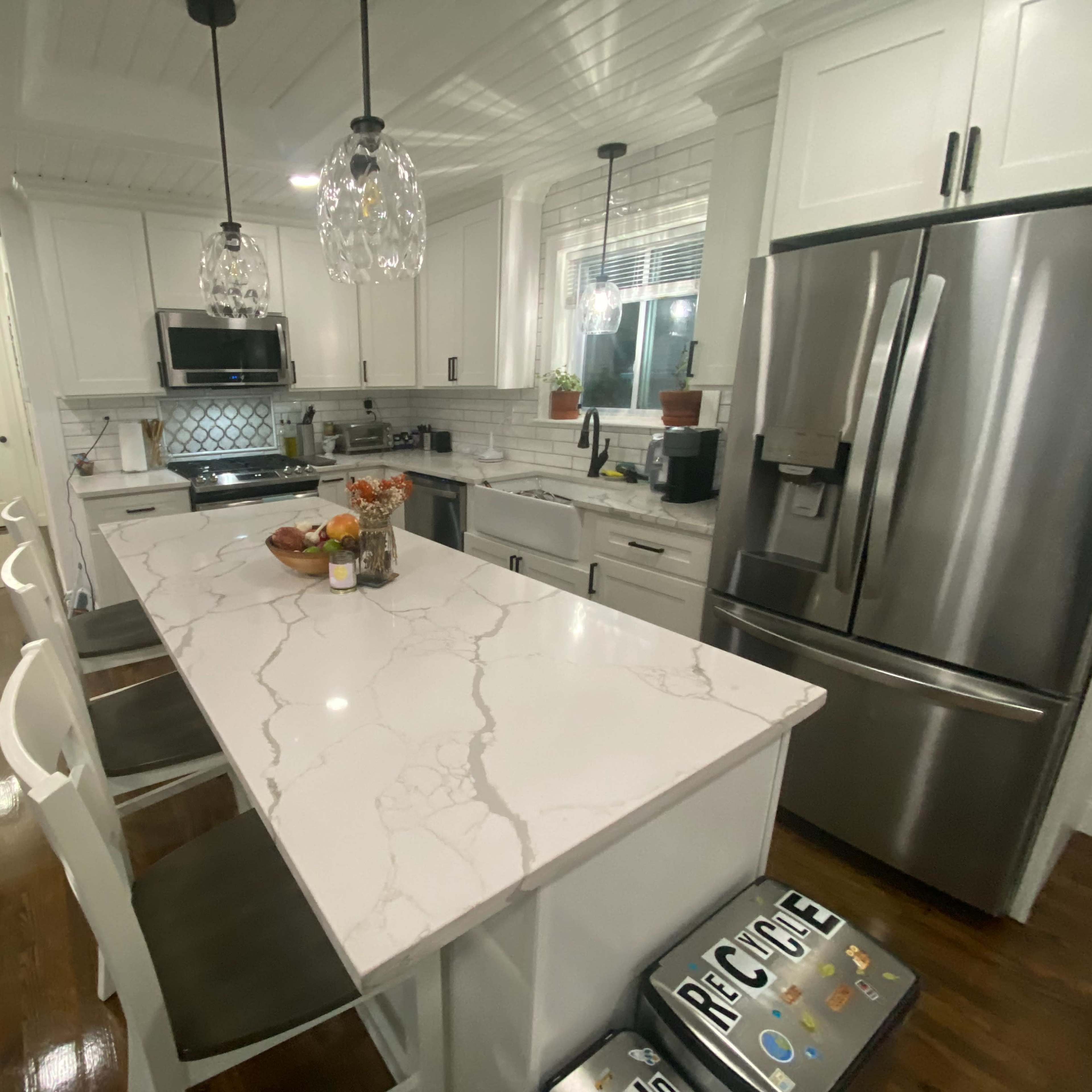 A modern kitchen features white cabinetry, a large marble island with seating, stainless steel appliances, and pendant lighting.