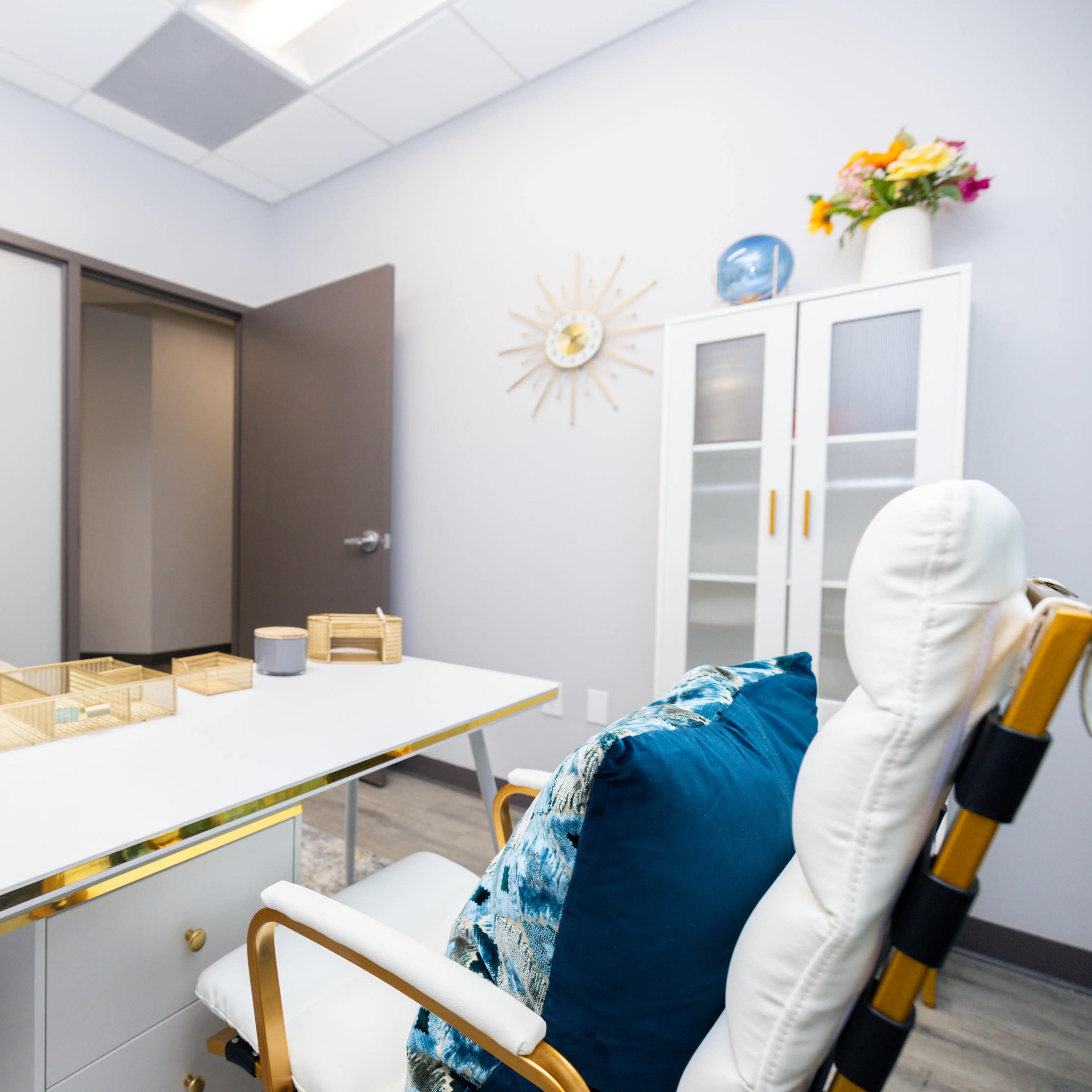 A modern office interior with a white desk, a chair, decorative plants, and a light-colored wall adorned with wall art and a cabinet.