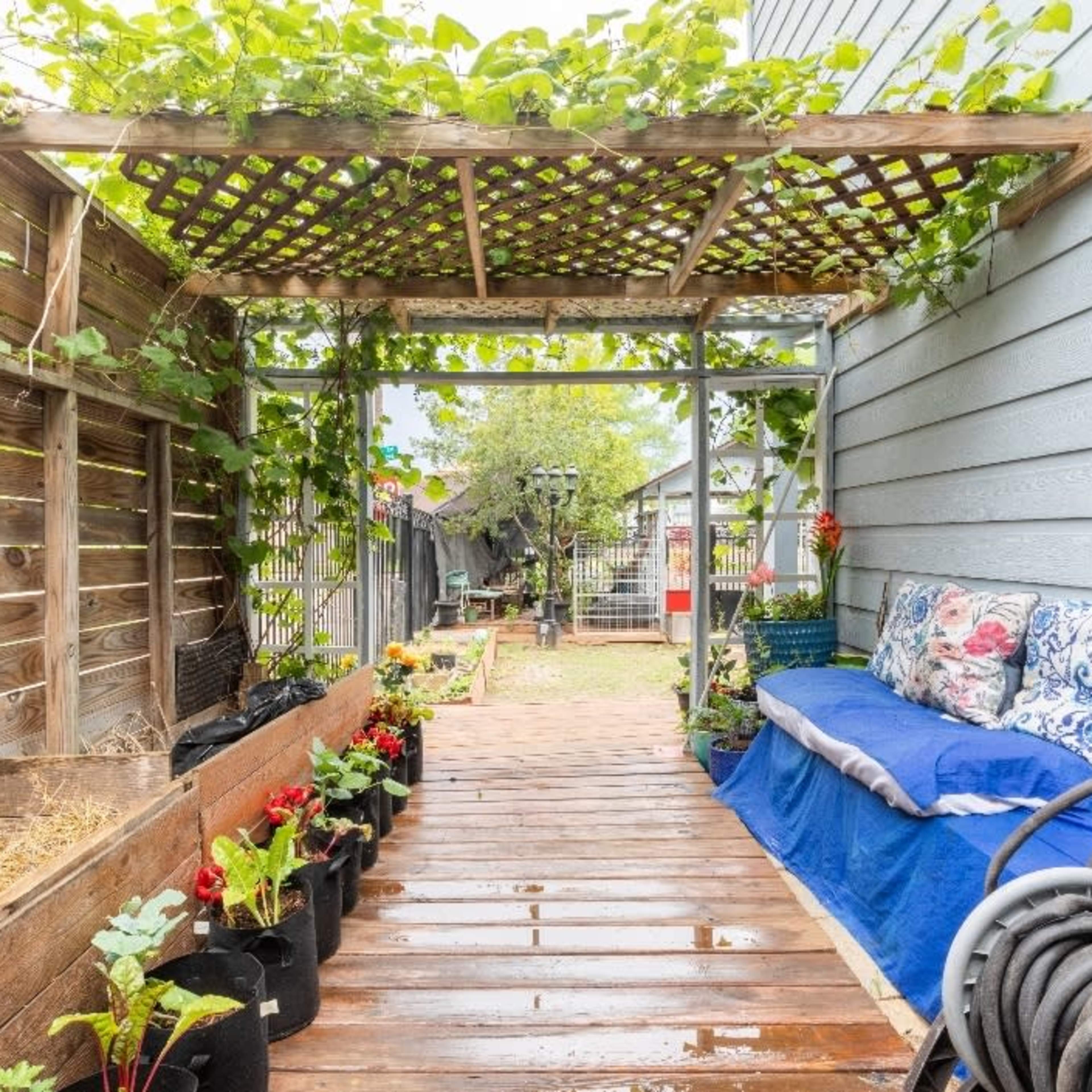 A wooden pathway leading through a garden area with potted plants on either side and a trellis overhead covered in greenery.