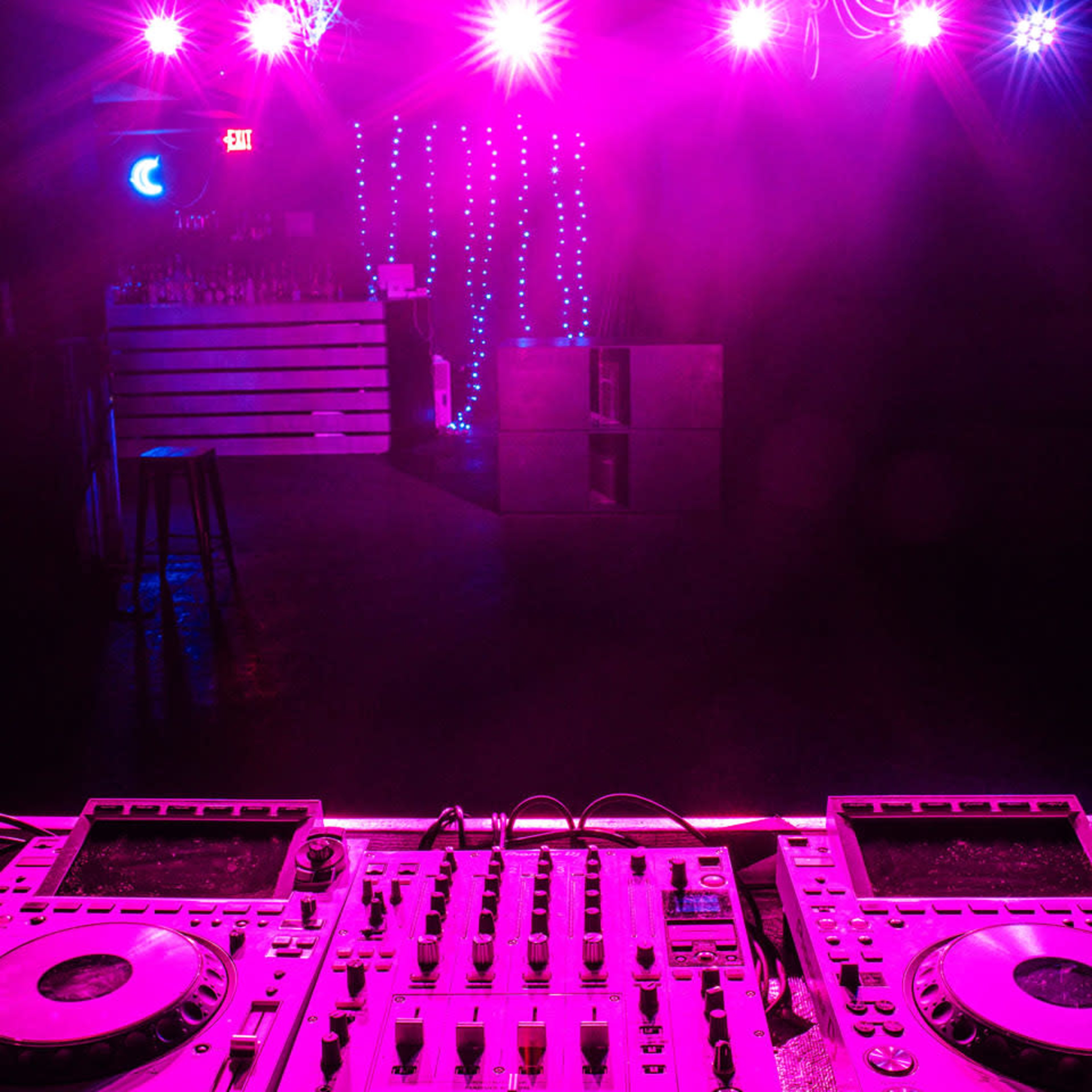 A dimly lit nightclub scene with a DJ setup featuring turntables and mixers in the foreground, illuminated by pink and blue lights.