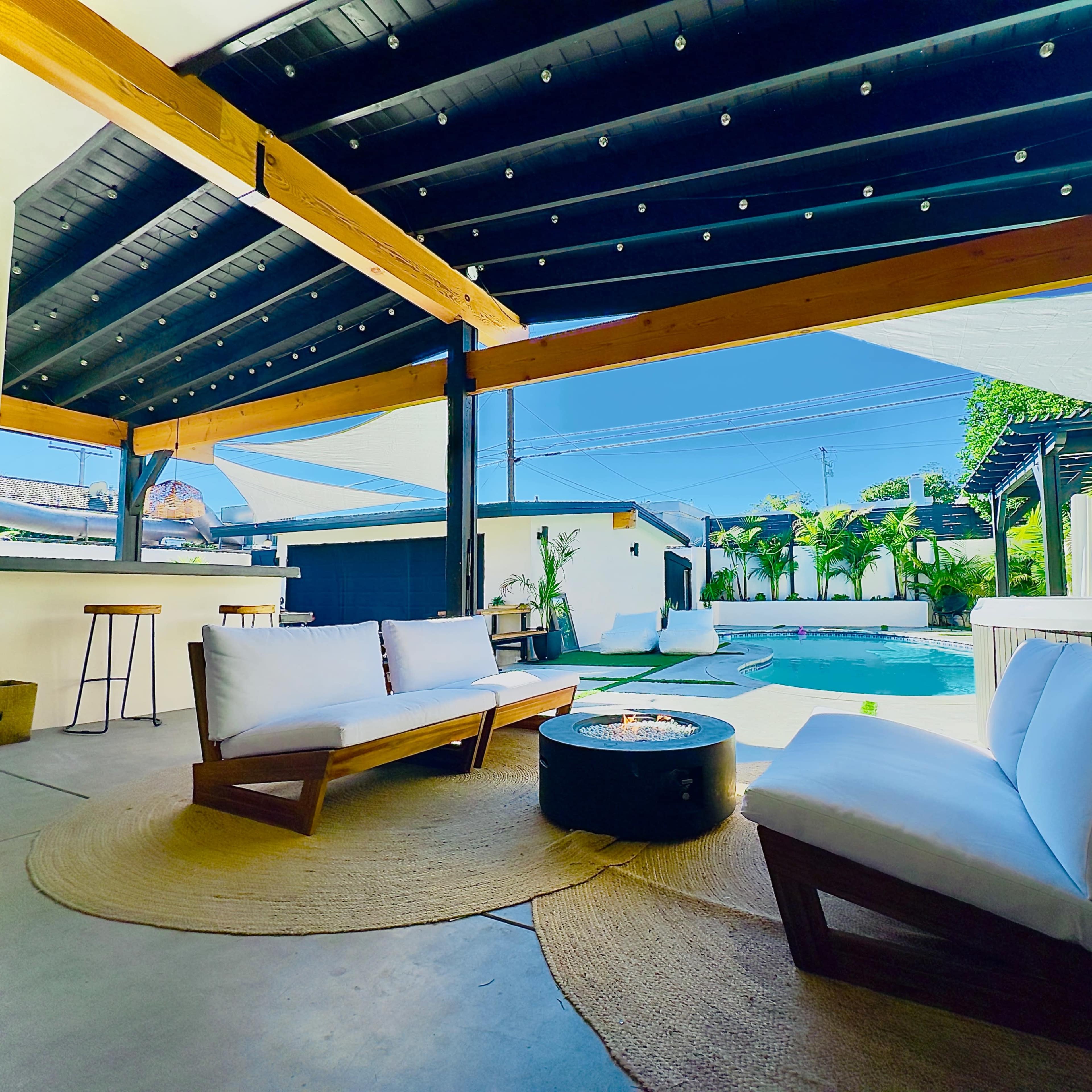 A covered patio area with two white sofas arranged around a circular fire pit, overlooking a swimming pool and landscaped yard.
