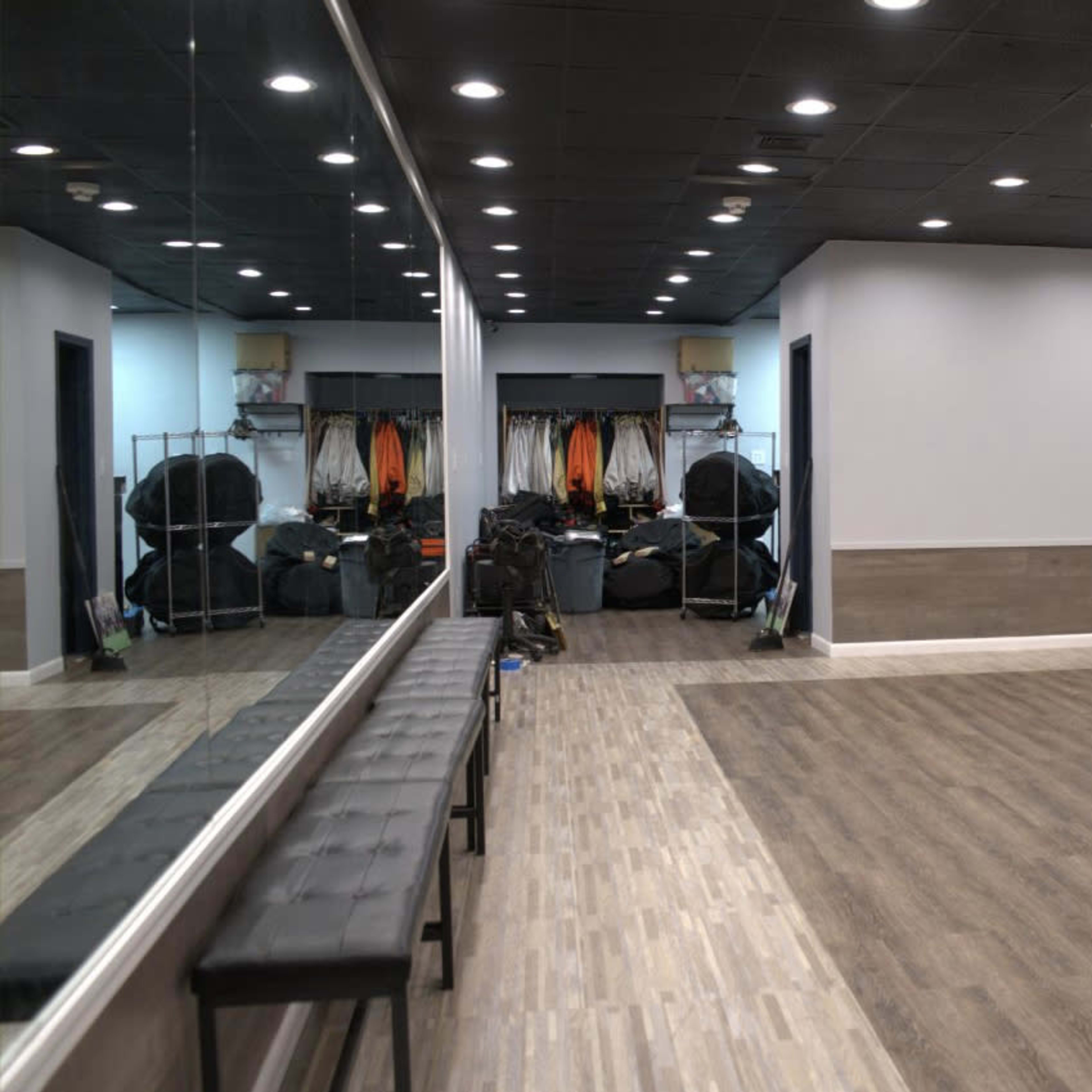 A spacious, well-lit dance studio with a mirrored wall and a bench along one side, featuring neatly arranged equipment in the background.