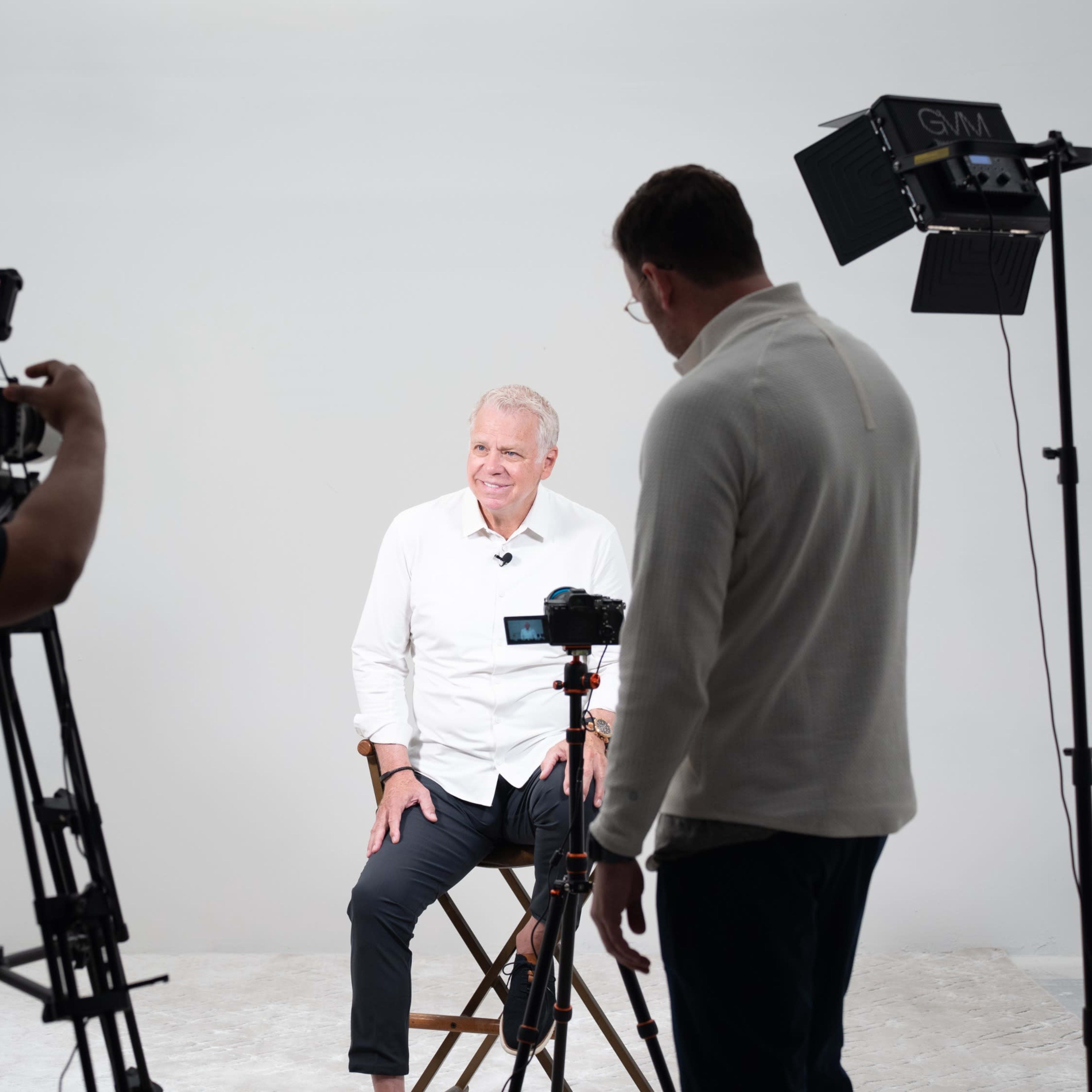 A man sits on a stool in a studio setting while being filmed by a crew using multiple cameras and lights.