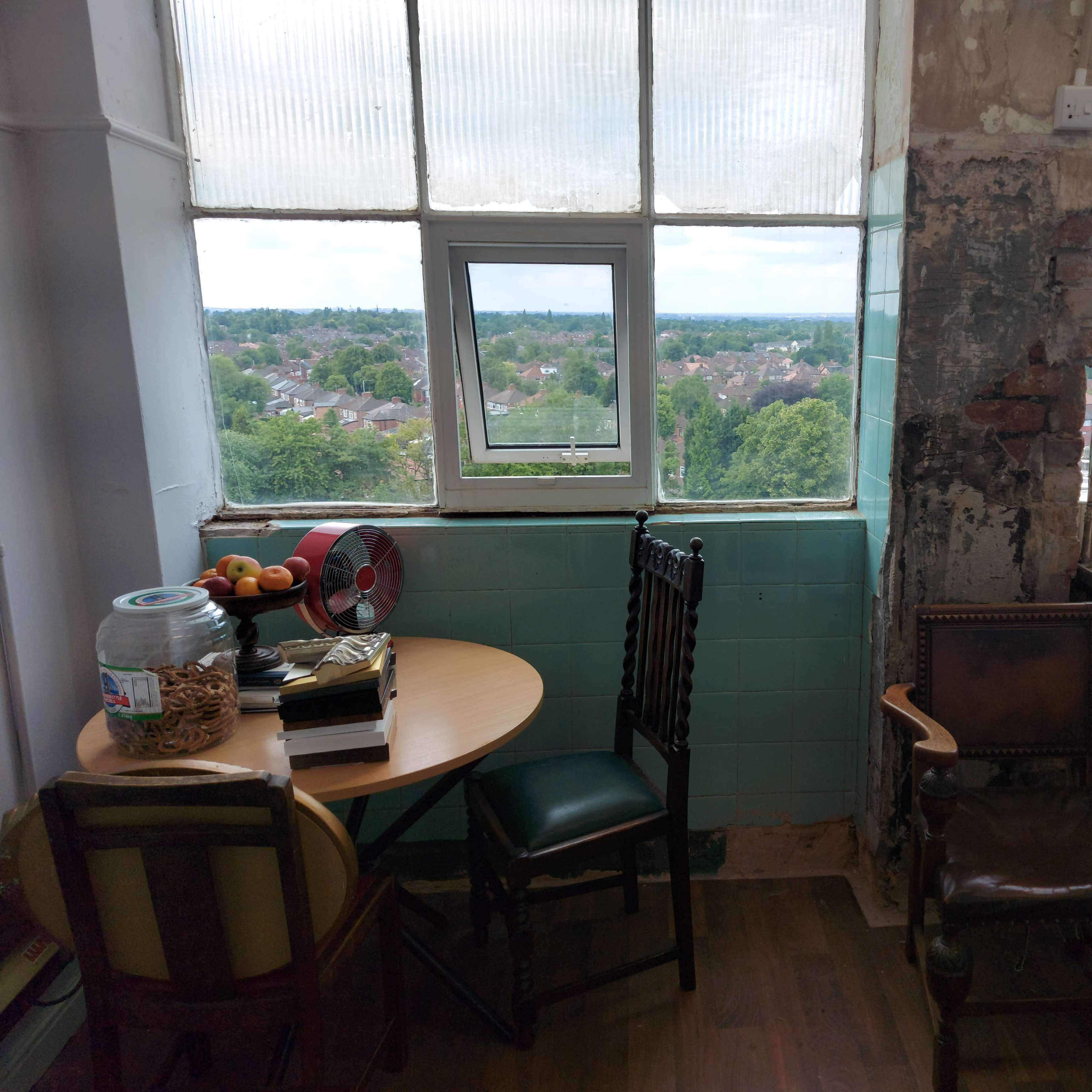 A small room features a table with fruit and a fan, two chairs, a kitchen area, and large windows providing a view of the surrounding landscape.