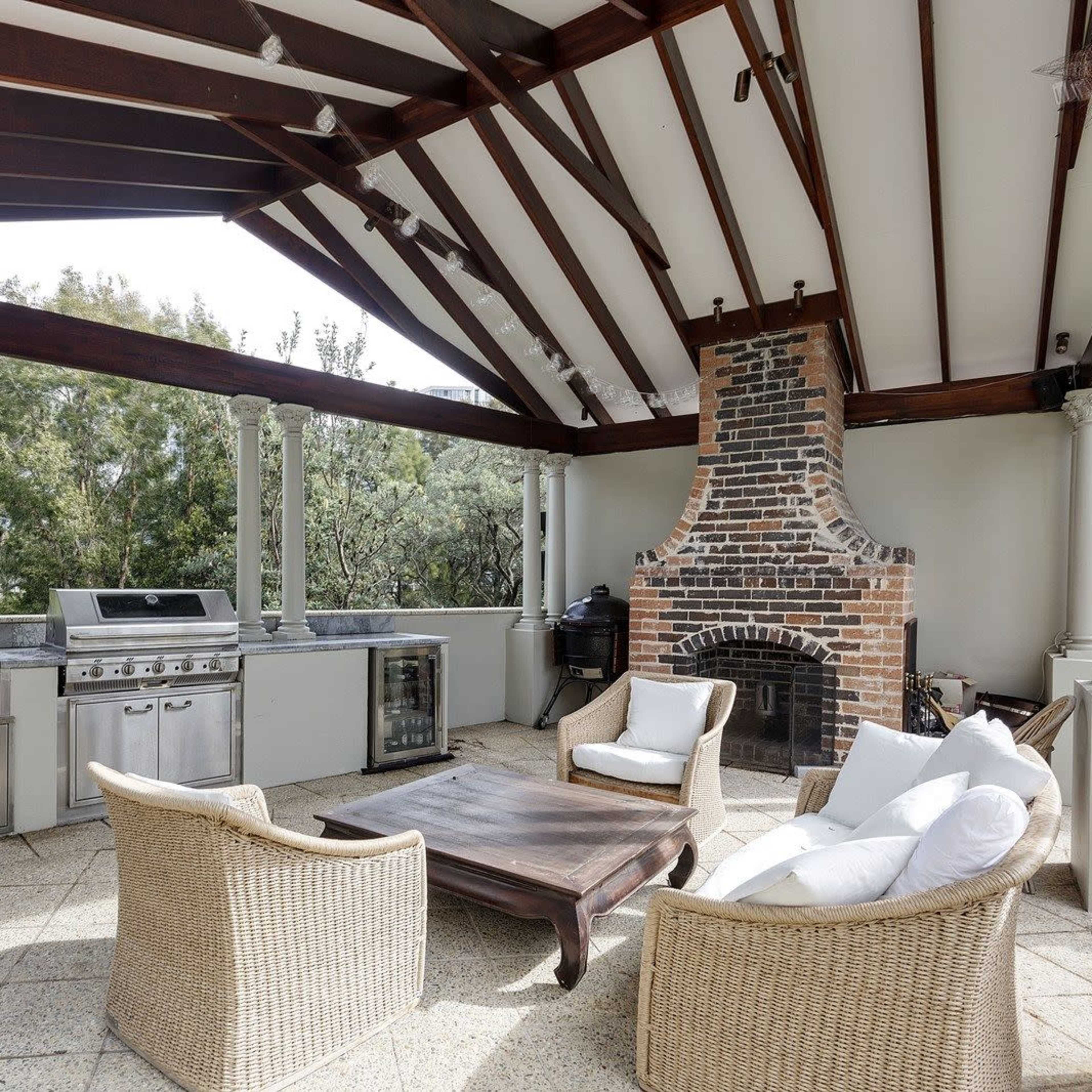 An outdoor kitchen with a grill, a stone fireplace, and seating arranged under a wooden pergola.