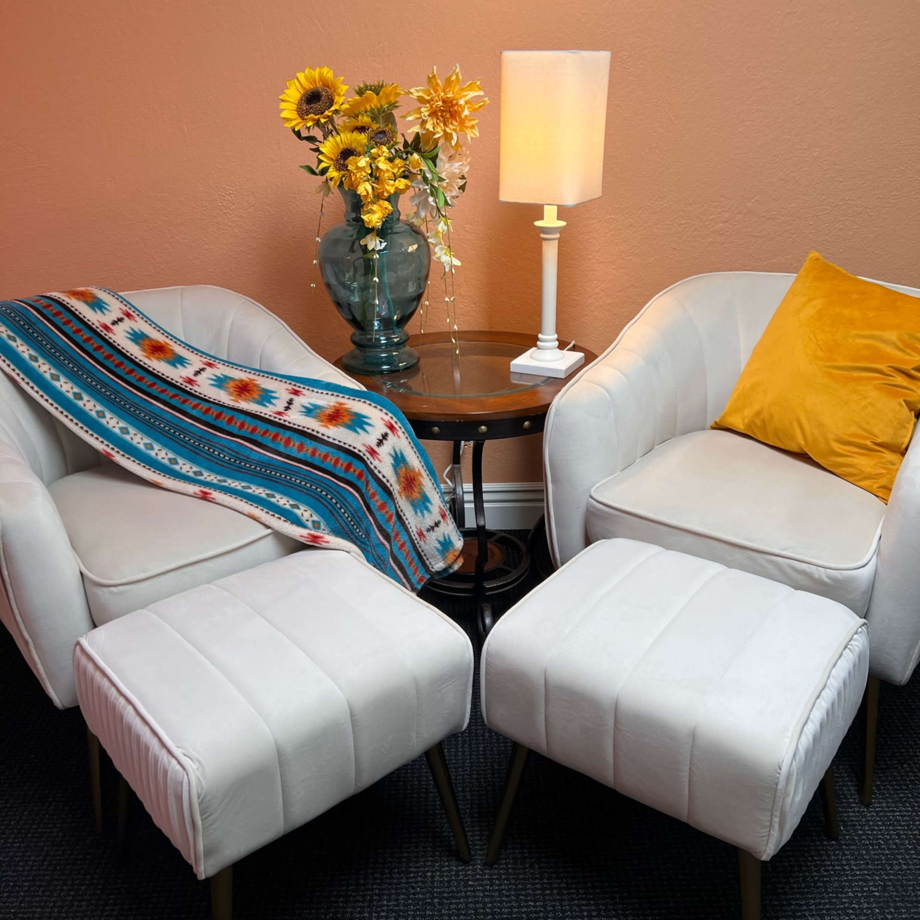 Two white armchairs are positioned alongside a small round table that holds a vase of sunflowers and a lamp, with a colorful blanket draped over one chair and a yellow cushion on another.