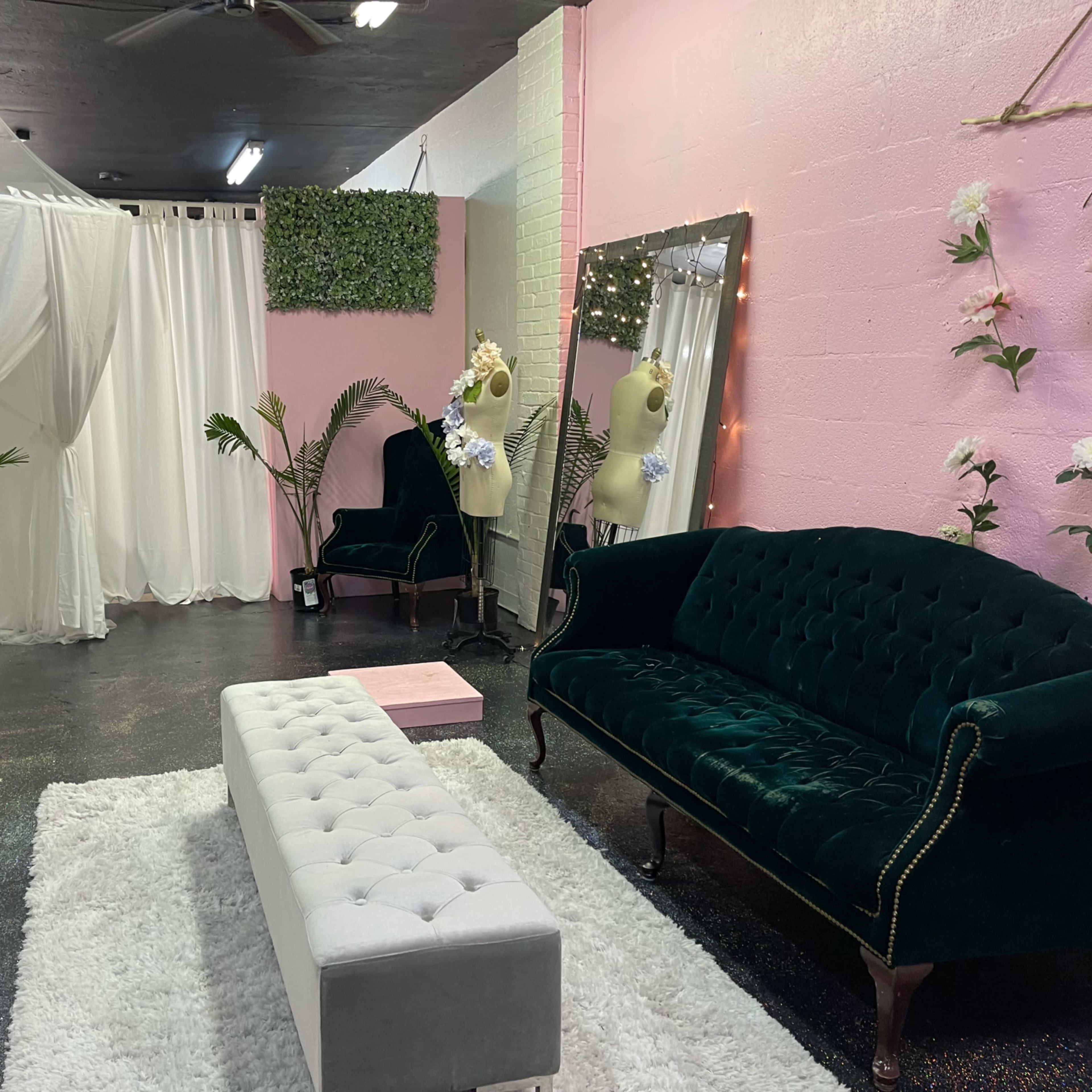 A room featuring a green velvet sofa, a white bench, and draped fabric partitions, with decorative plants and a large mirror.