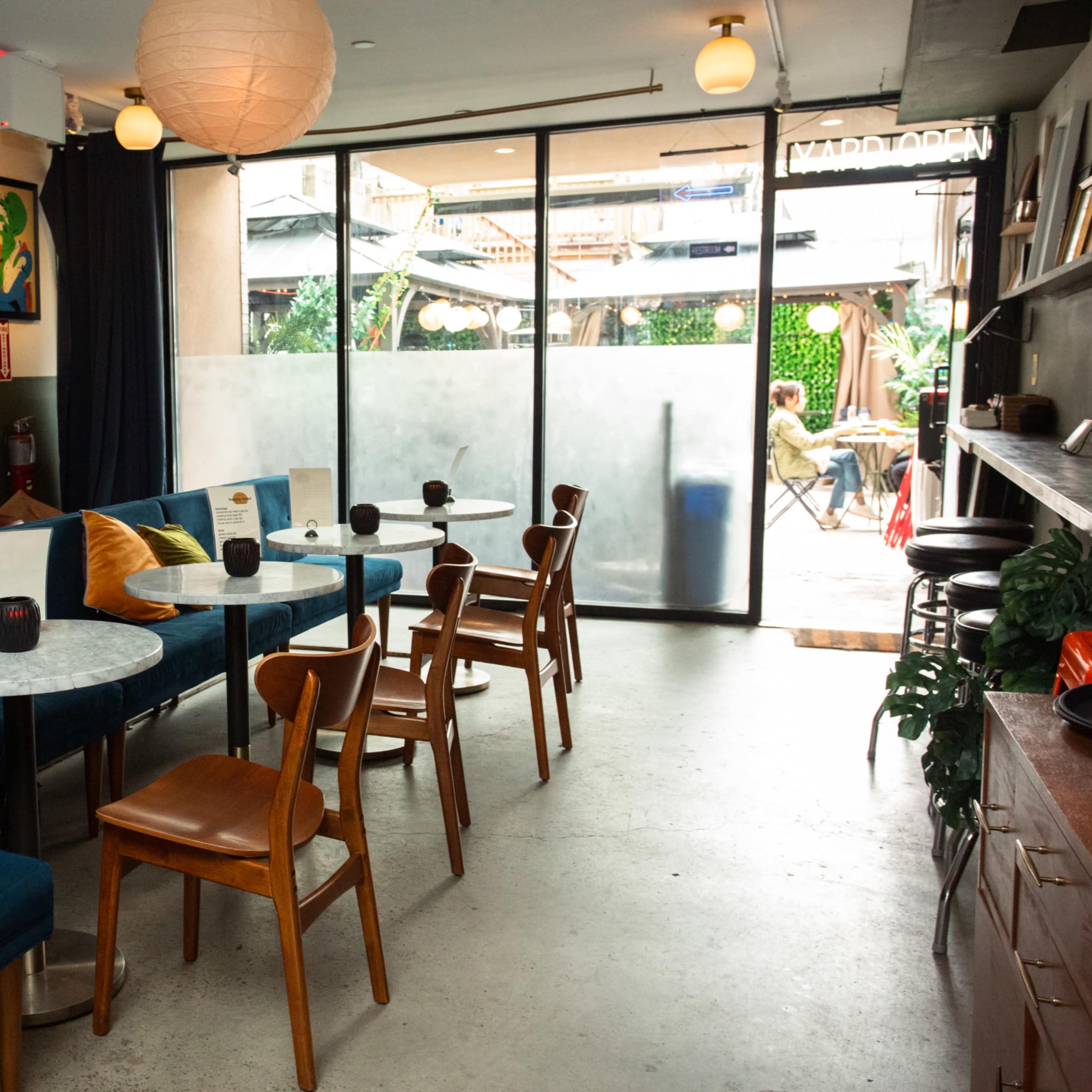 A cozy cafe interior with blue velvet seating, wooden chairs, and a view of an outdoor seating area through large glass doors.